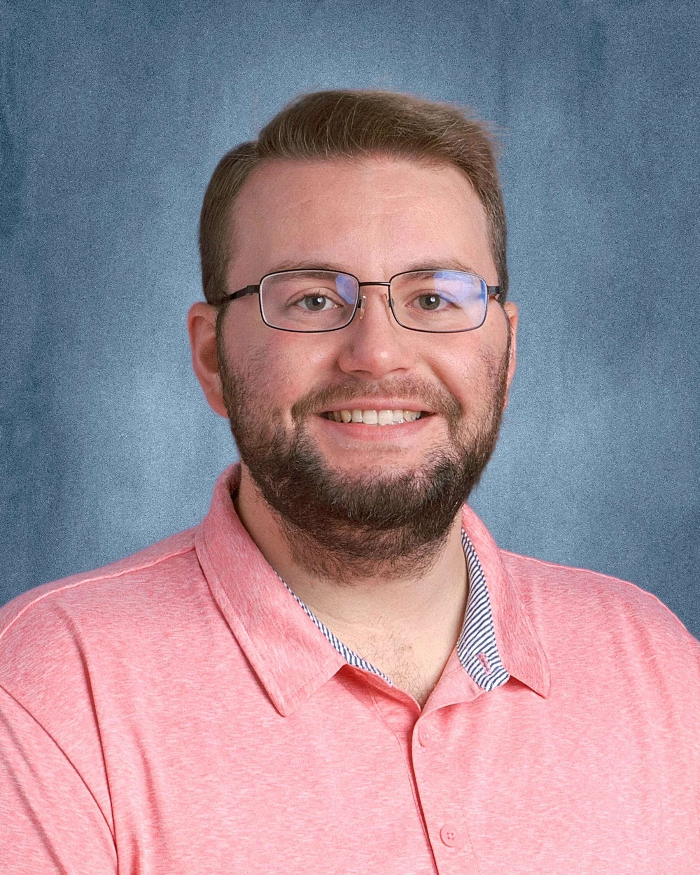 A close-up portrait of a smiling man with glasses, light skin, short brown hair, wearing a pink collared shirt with a striped inner lining, set against a plain blue background.