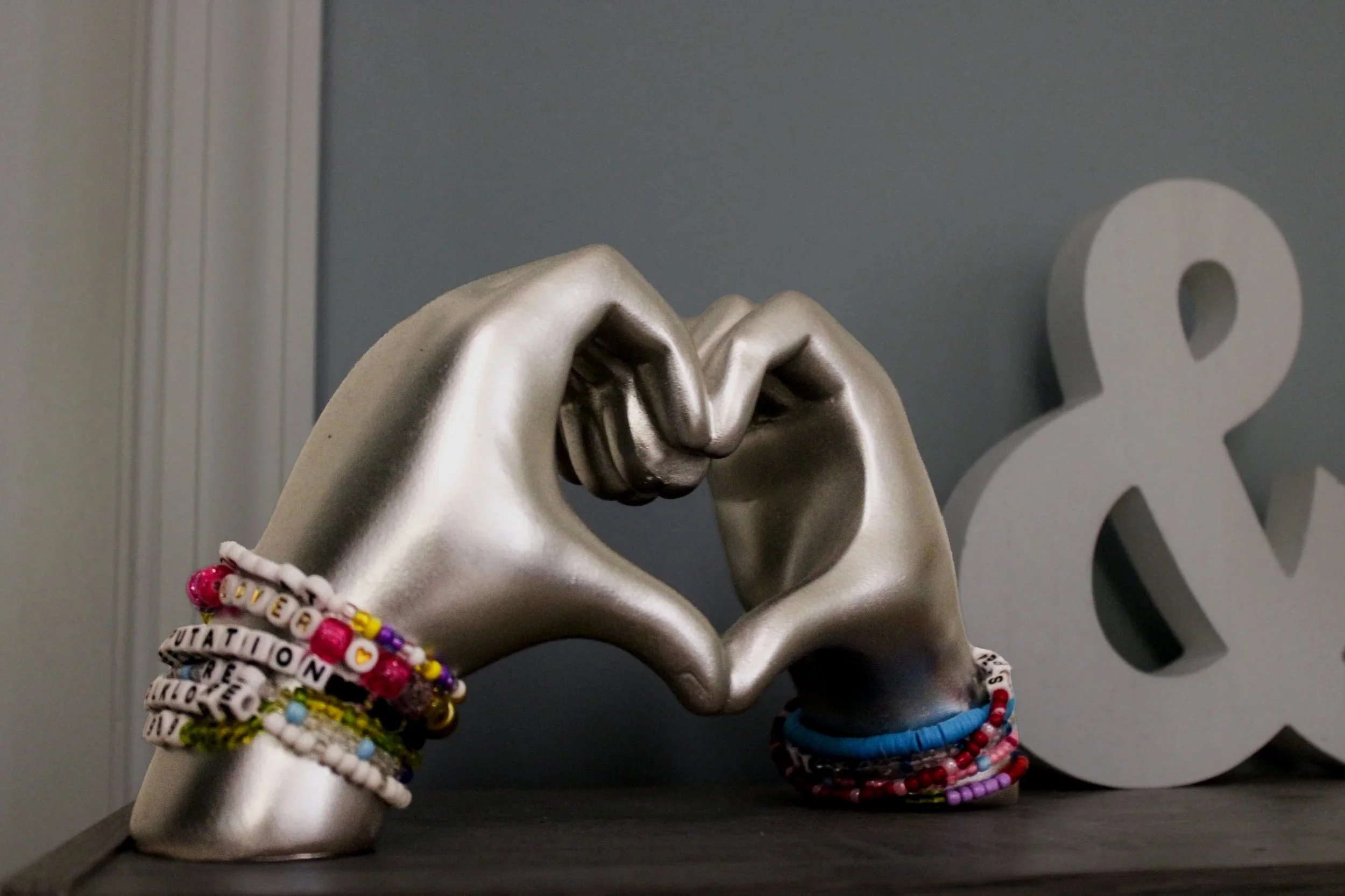 A metallic hand sculpture in a fist pose, decorated with colorful beaded bracelets with words and symbols. In the background, a large number '&' is visible, set against a gray wall.