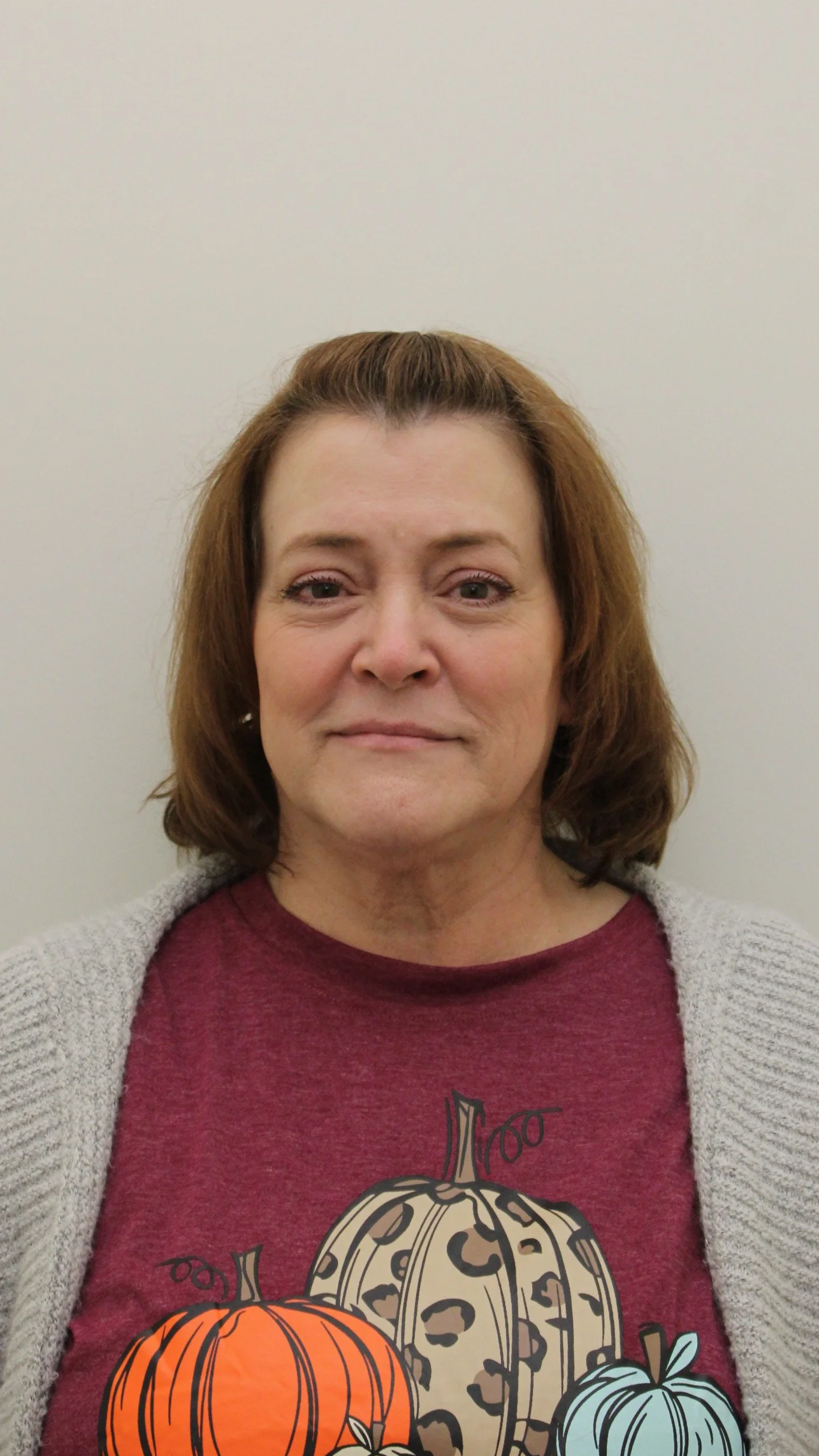 A woman with shoulder-length brown hair, wearing a maroon shirt with pumpkin illustrations and a gray cardigan, standing against a plain white wall.