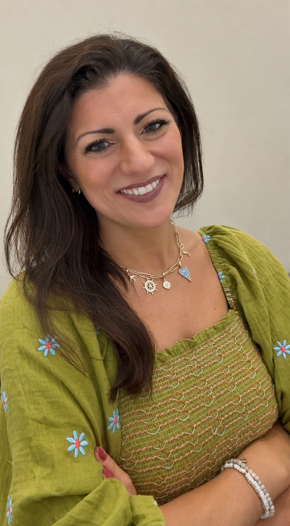 A woman with long dark hair, wearing a green embroidered top and a matching green jacket with blue flower embroidery, smiling with her arms crossed, accessorized with a layered necklace featuring nautical charms and a blue heart, and a white beaded bracelet.