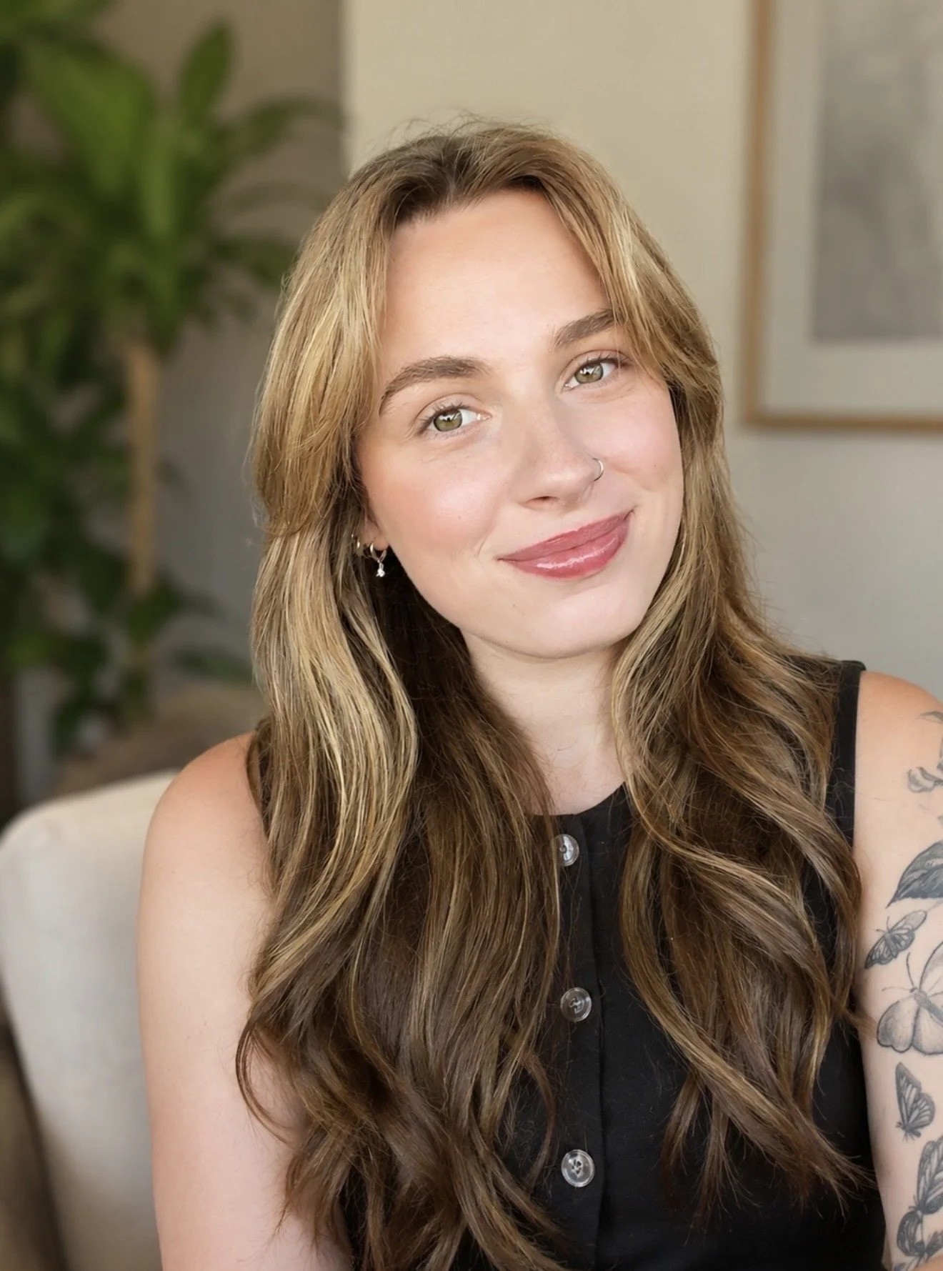 A young woman with long, wavy light brown hair, light skin, and a nose ring, smiling while sitting on a beige couch. She has tattoos on her left arm and is wearing earrings and a sleeveless black top with buttons. In the background, there is a leafy green plant on the left and a framed picture on the wall.