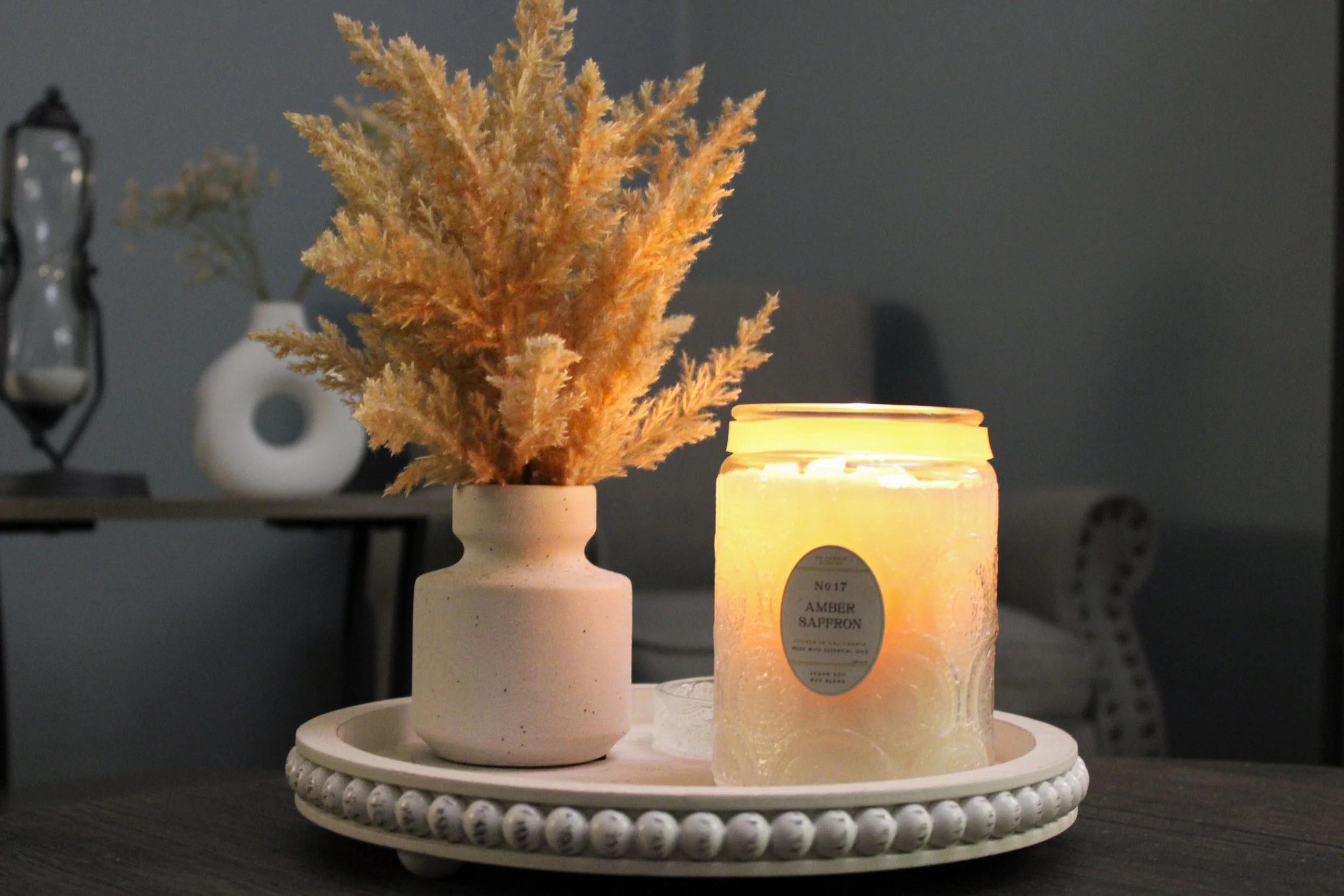 A decorative arrangement with dried beige pampas grass in a white vase, a lit cream-colored candle labeled "Amber Saffron", and a round tray with beaded edges, on a dark surface in a dimly lit room.