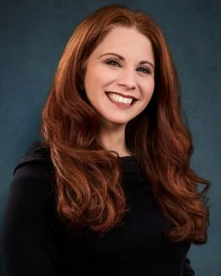A woman with long red hair smiling against a dark teal background.