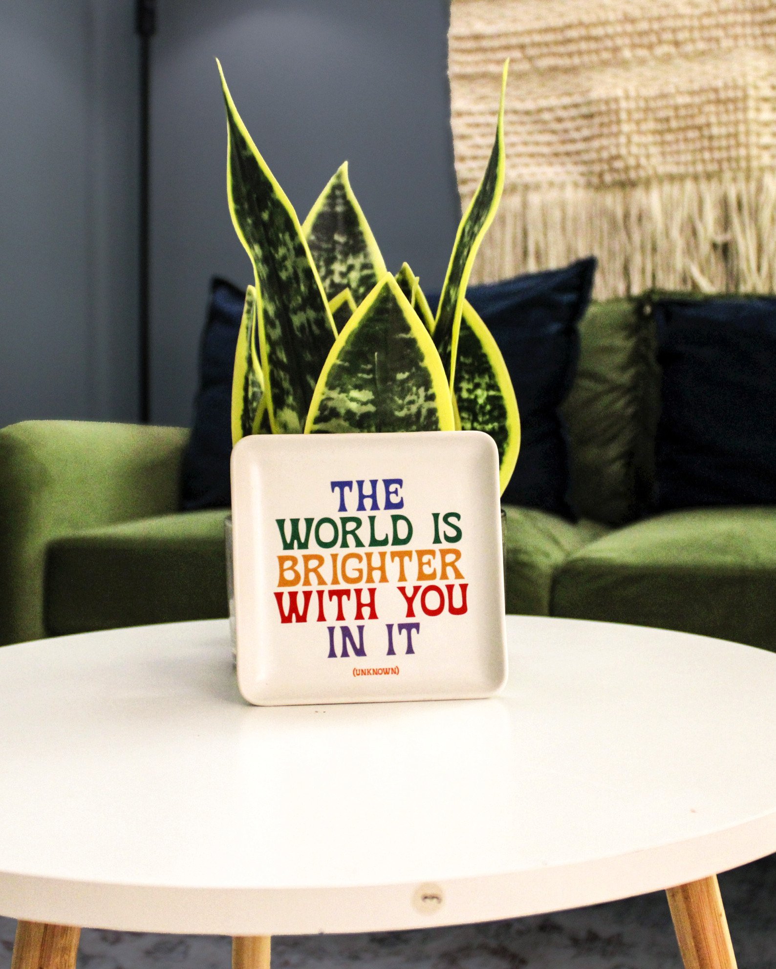 A decorative sign with the message "The world is brighter with you in it" placed on a white round table. Behind the table is a plant with pointed, variegated green and yellow leaves, and two dark pillows on a couch.