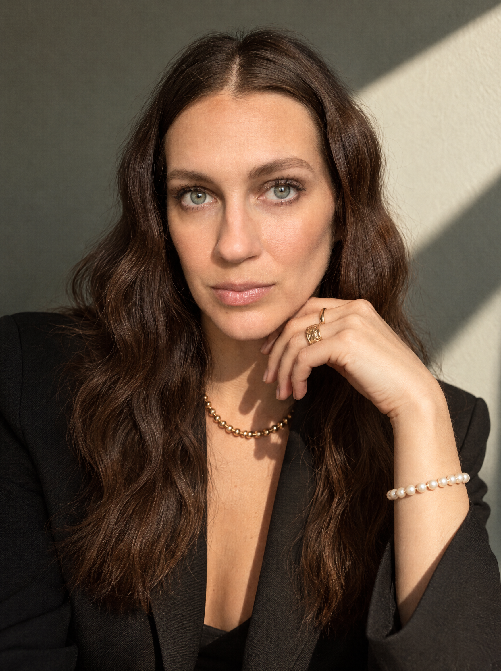 A woman with long wavy brown hair and green eyes, wearing a black blazer, pearl necklace and rings, looking directly at the camera, with her chin resting on her hand, in natural light.