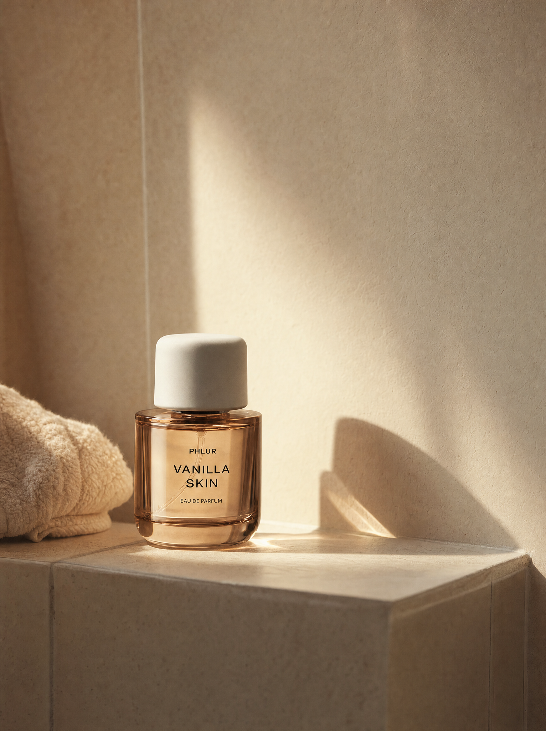A glass perfume bottle labeled "Phlur Vanilla Skin Eau de Parfum" sitting on a beige tile surface with a beige textured wall in the background. There is a rolled towel partially visible on the left side of the image.