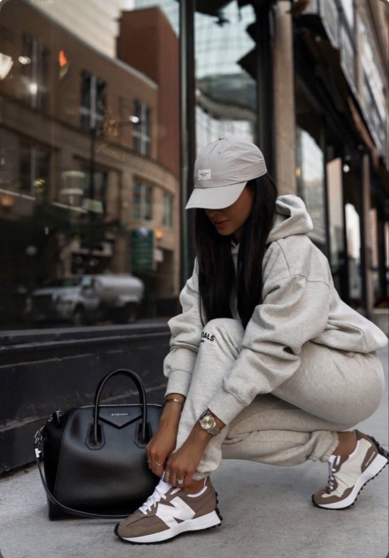 A woman dressed in a light-colored hoodie, sweatpants, and sneakers, wearing a cap and a watch, is squatting on the sidewalk beside a black handbag, in an urban setting with buildings reflected in the window behind her.