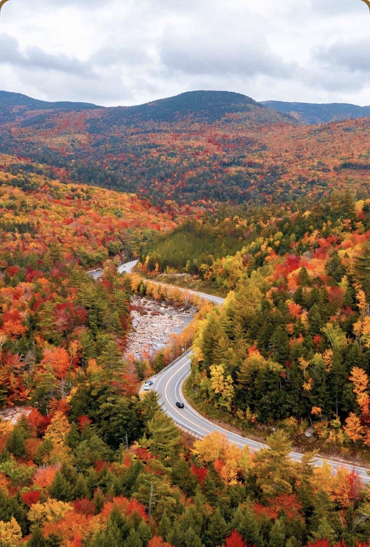 A winding mountain road through a colorful autumn forest with red, orange, yellow, and green leaves, and mountains in the background under cloudy skies.
