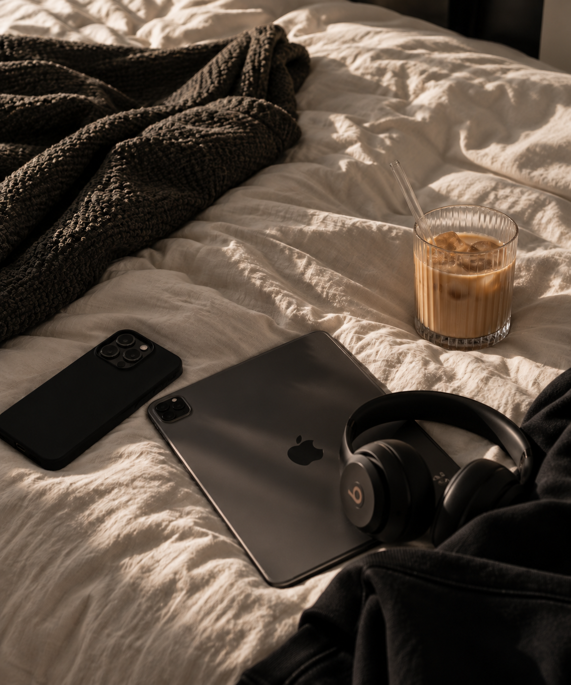 A cozy bed with a dark knitted blanket, an iPad, two iPhones, a glass of iced coffee, and black Beats headphones.