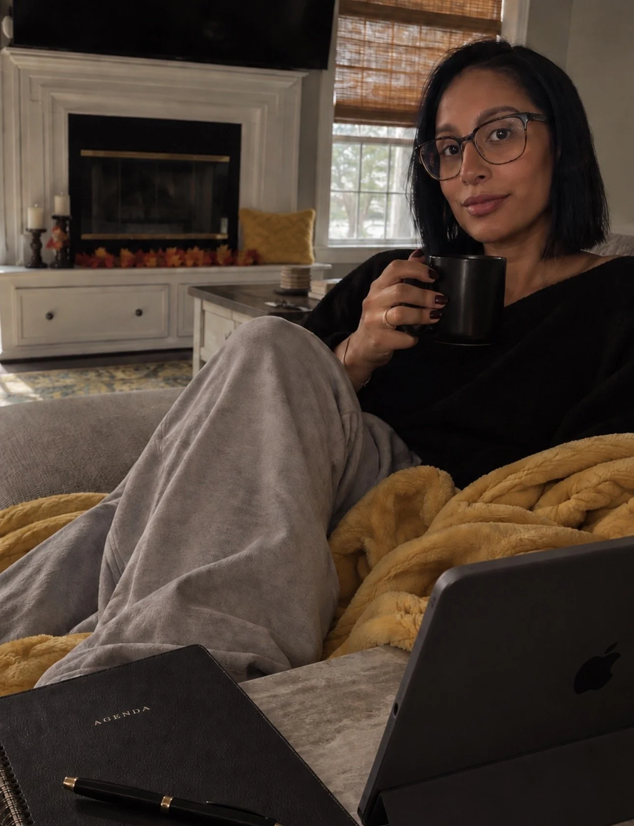 A woman with black hair and glasses sitting on a couch, holding a black mug, with a yellow blanket on her lap, in a cozy living room with a fireplace, window, and various decor.