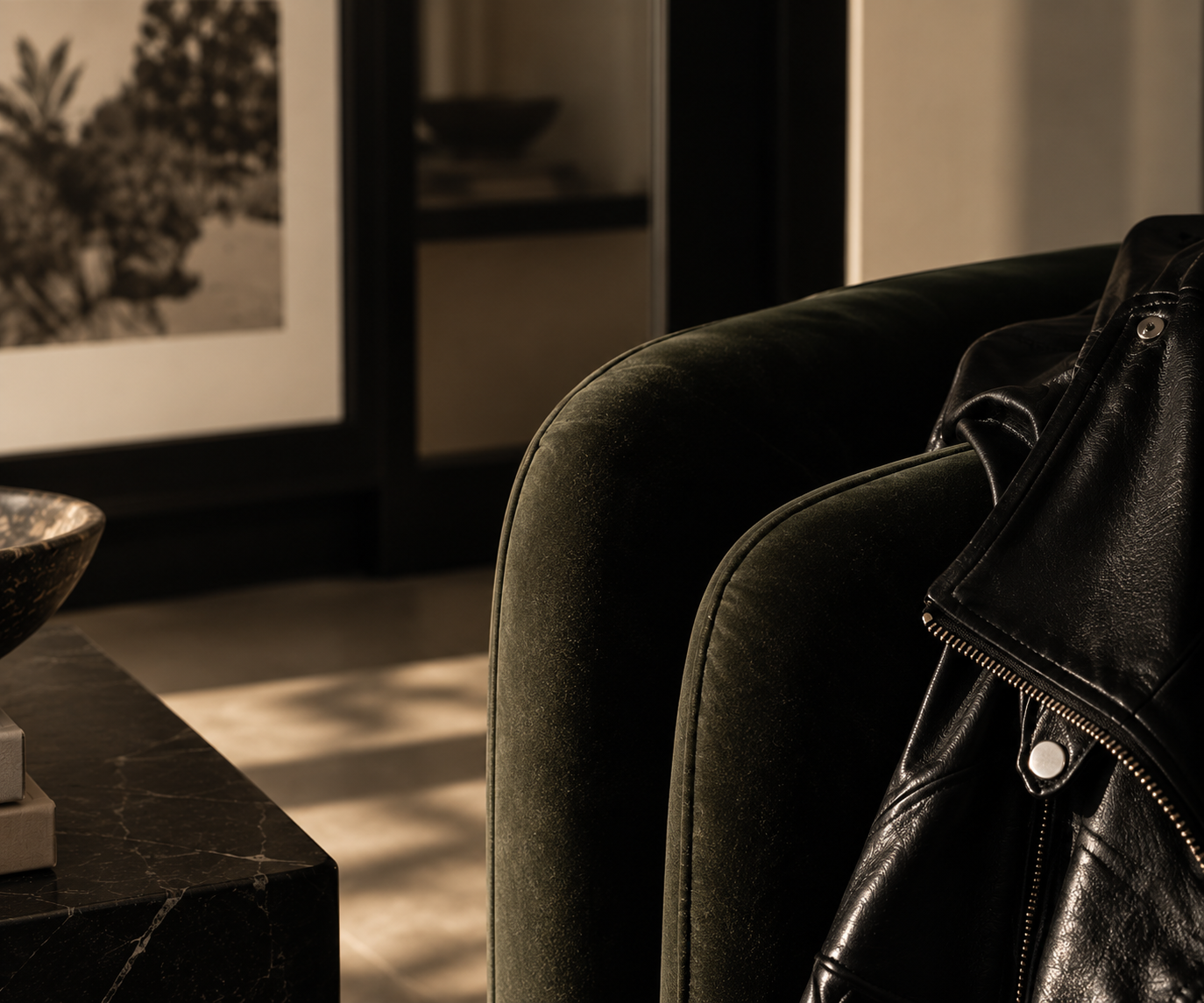 Close-up of a black leather jacket draped over the green velvet arm of a sofa in a dimly lit room.