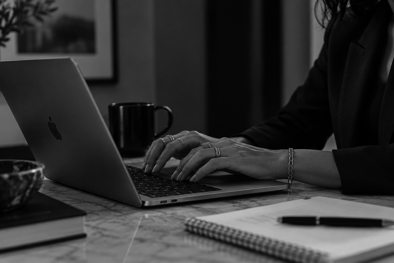 A person working on a laptop at a desk with notebooks, a pen, a coffee mug, and a bowl in black and white.