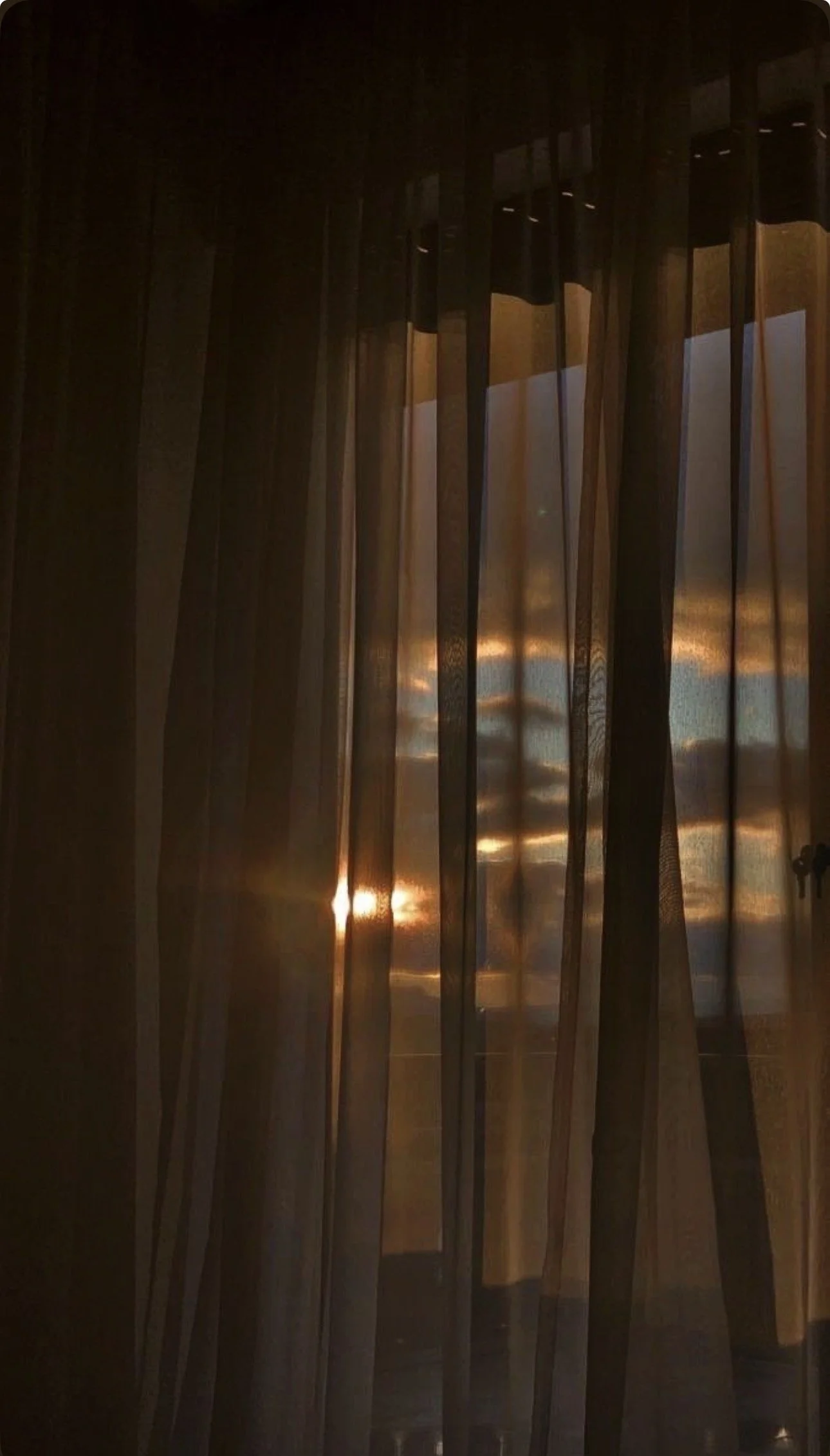 Sunset view through a window covered by dark curtains, creating silhouettes and reflections of the sunset colors.