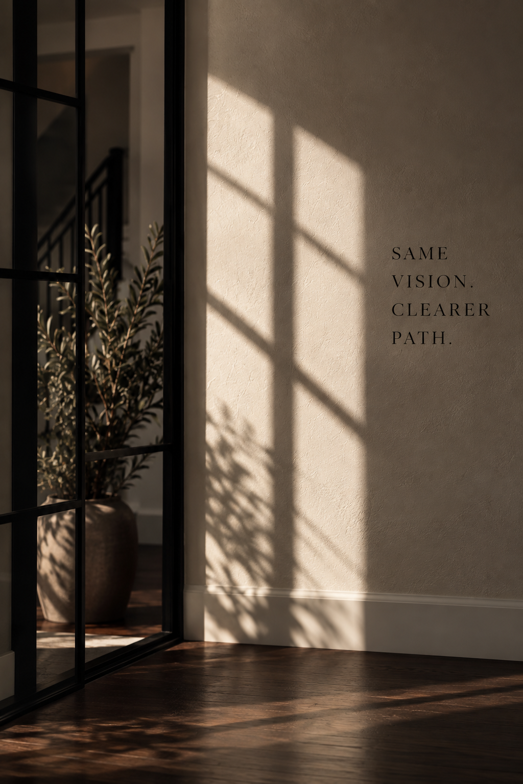 Sunlight casting shadows of window panes onto a beige wall, with black lettering that reads 'SAME VISION. CLEARER PATH.', next to a glass door and a potted plant inside.