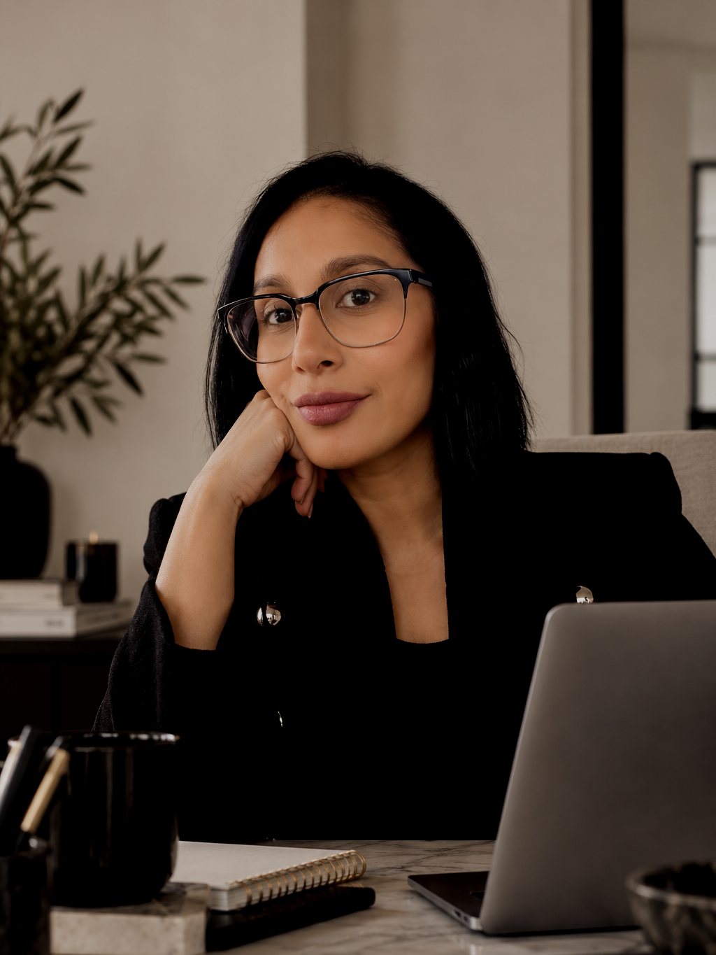 A woman with black hair wearing glasses sits at a desk, resting her chin on her hand, looking at the camera with a slight smile. The desk has a laptop, a notebook, and office supplies.