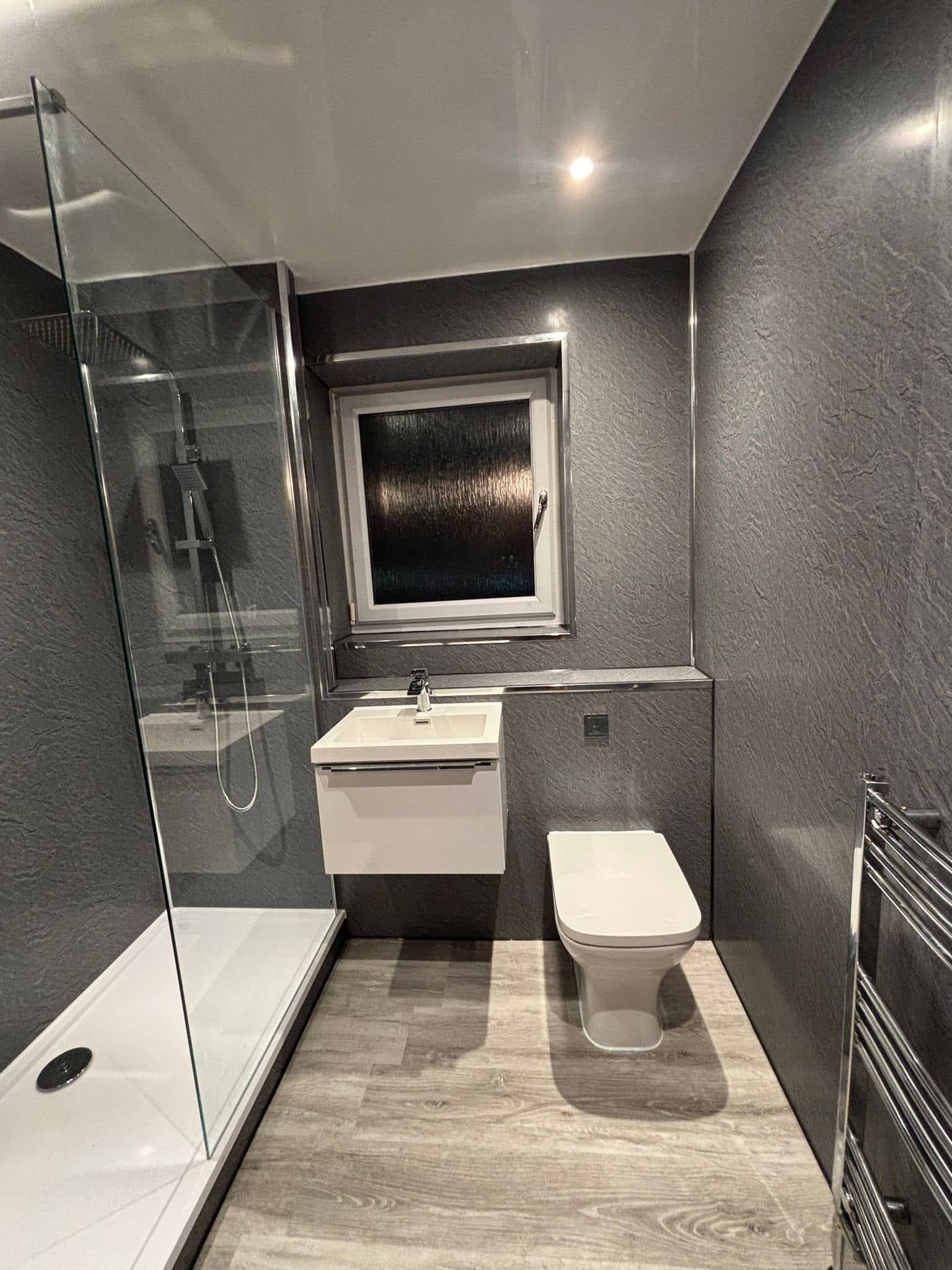 Modern bathroom with dark textured walls, a glass shower enclosure with a rainfall showerhead, a small white sink with a cabinet, a window above the sink, a wall-mounted toilet, and a heated towel rack.