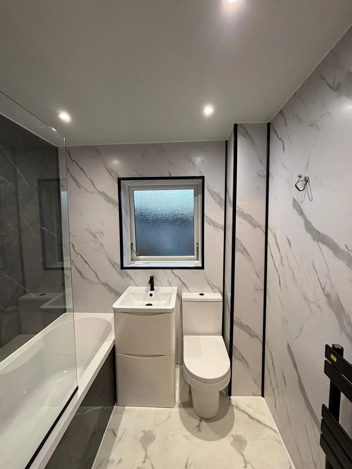 Modern bathroom with white marble walls, small window, sink, toilet, and bathtub shower combination.