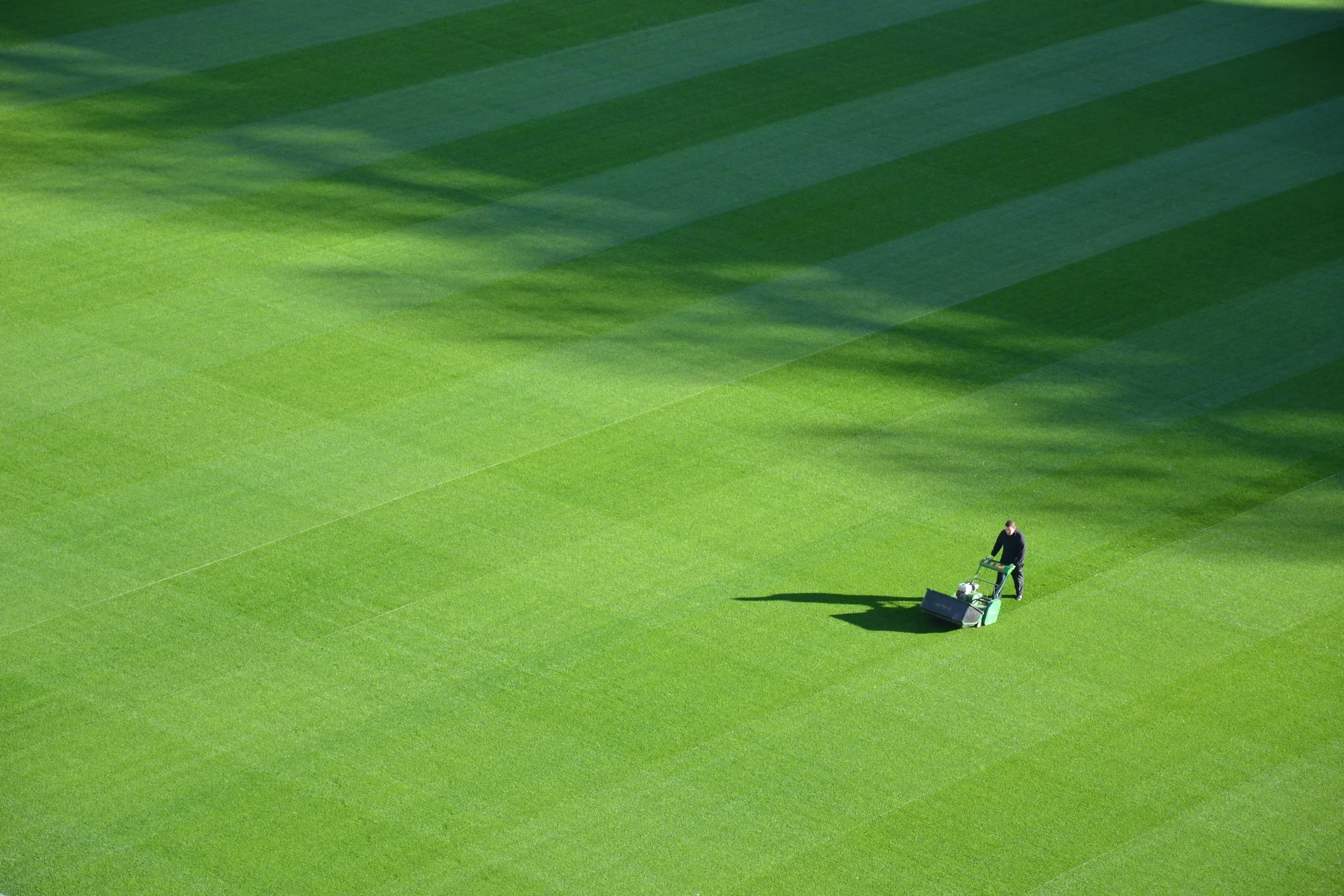 A person using a lawn mower to trim bright green grass on a large field, with long shadows cast by the sunlight.
