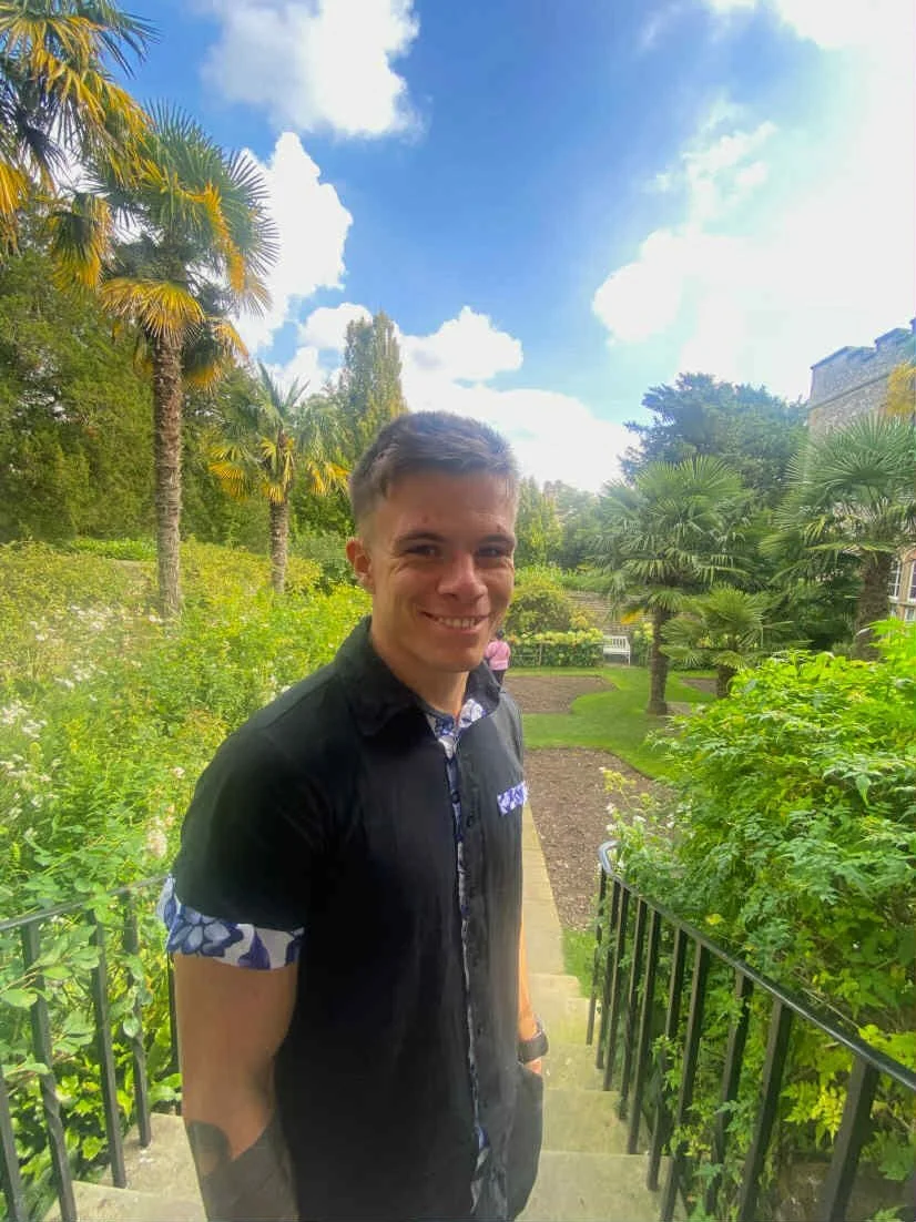 A young man smiling outdoors in a garden with lush green plants, trees, and a partly cloudy sky in the background.