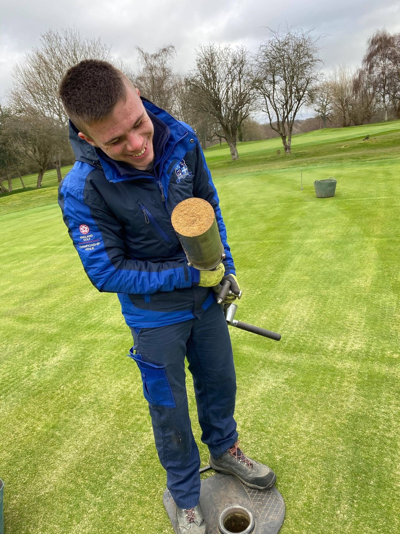 A man in a blue jacket and work boots is smiling and standing on a green golf course, holding a ground-penetrating tool over a hole in the ground.