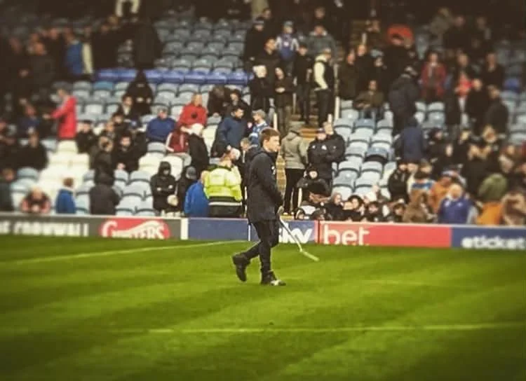 A person in dark clothing is walking across a football field while others stand or sit in the stands in the background.