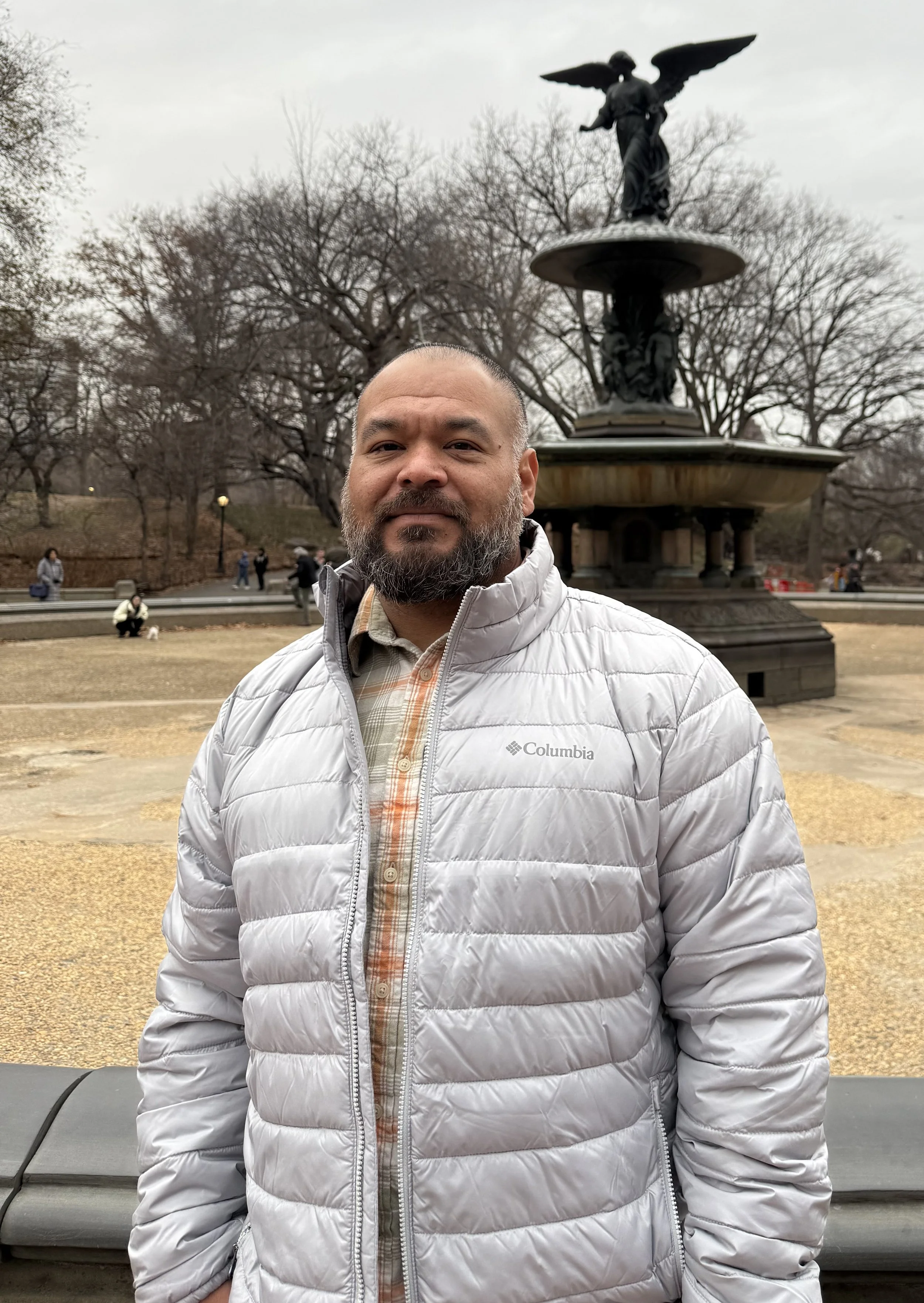 A man with a beard and short hair wearing a light gray puffy Columbia jacket and a plaid shirt stands outdoors in front of a large decorative fountain and leafless trees, on an overcast day.
