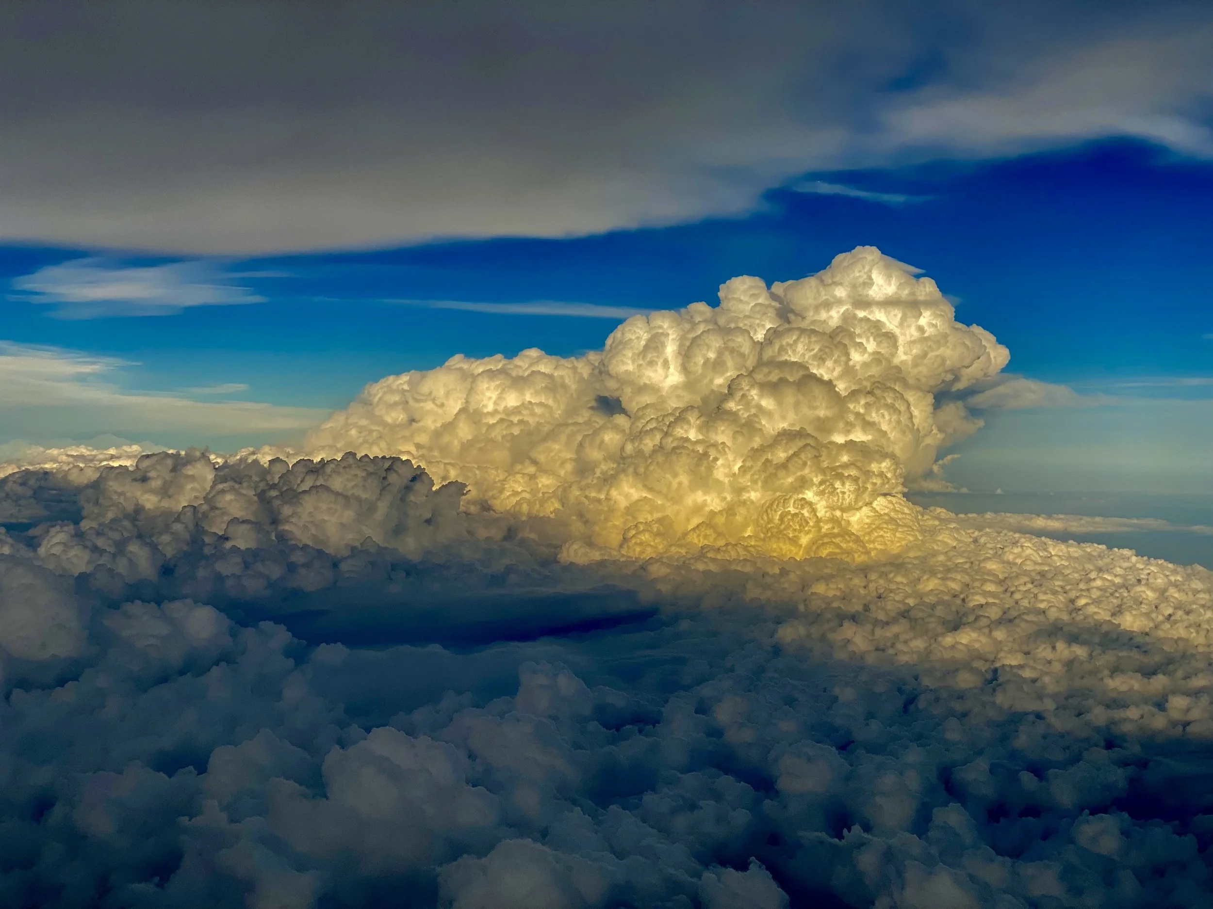 A view of a large cloud formation with a mix of bright white, yellowish-light, and dark gray clouds under a blue sky with some thinner clouds.