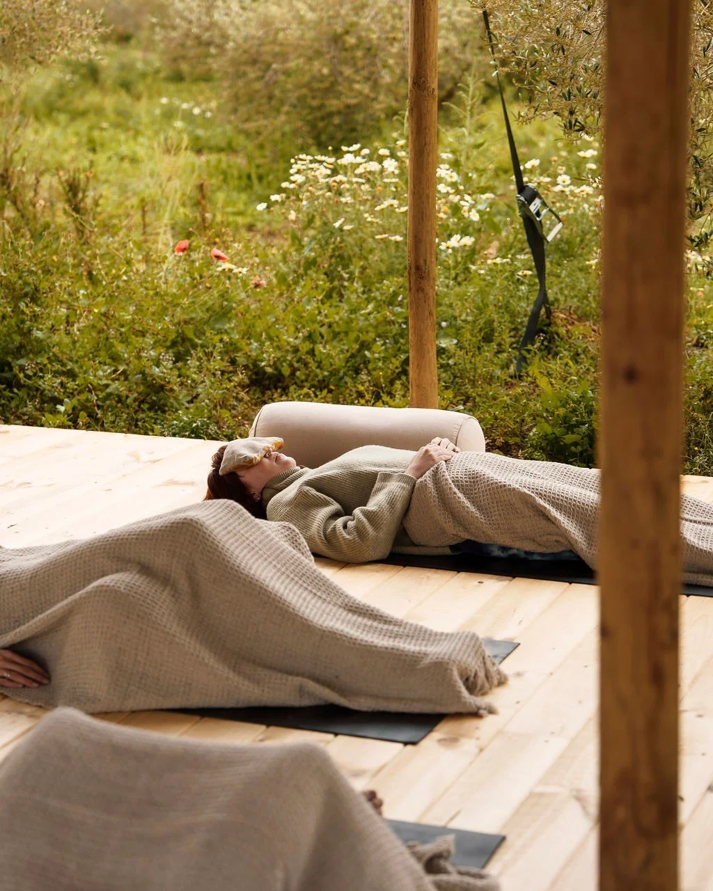 Guest resting under a waffle blanket with an eye pillow and bolster during a guided breathwork session on an outdoor deck among wildflowers in Mallorca