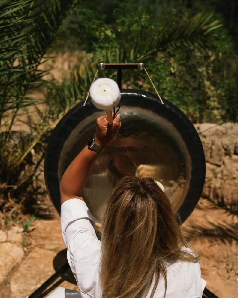 Sound practitioner playing a large gong in a Mallorcan garden among palms and stone walls, during a private sound bath.