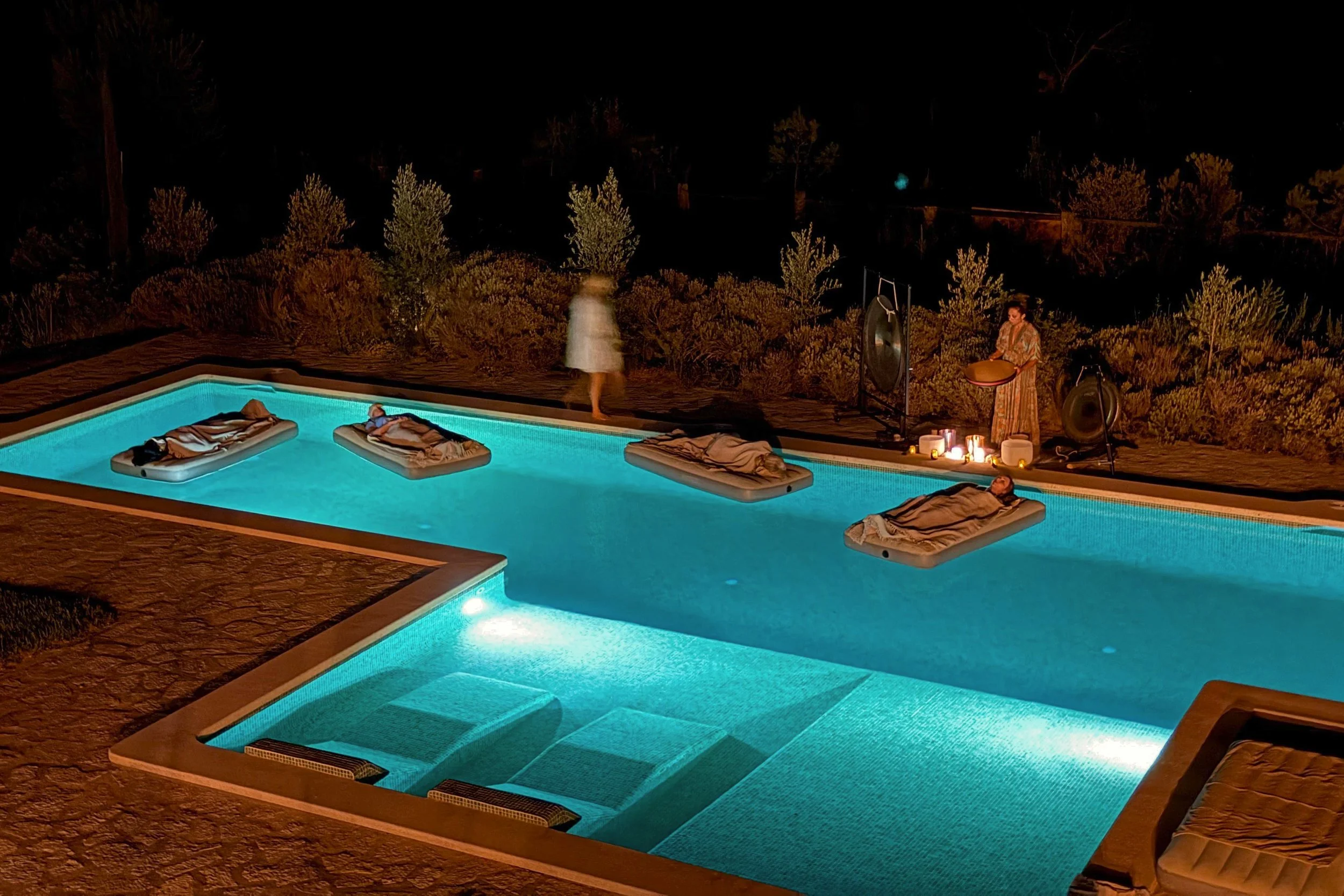 Guests on floating beds in a lit villa pool at night while a practitioner plays live sound instruments, Mallorca.
