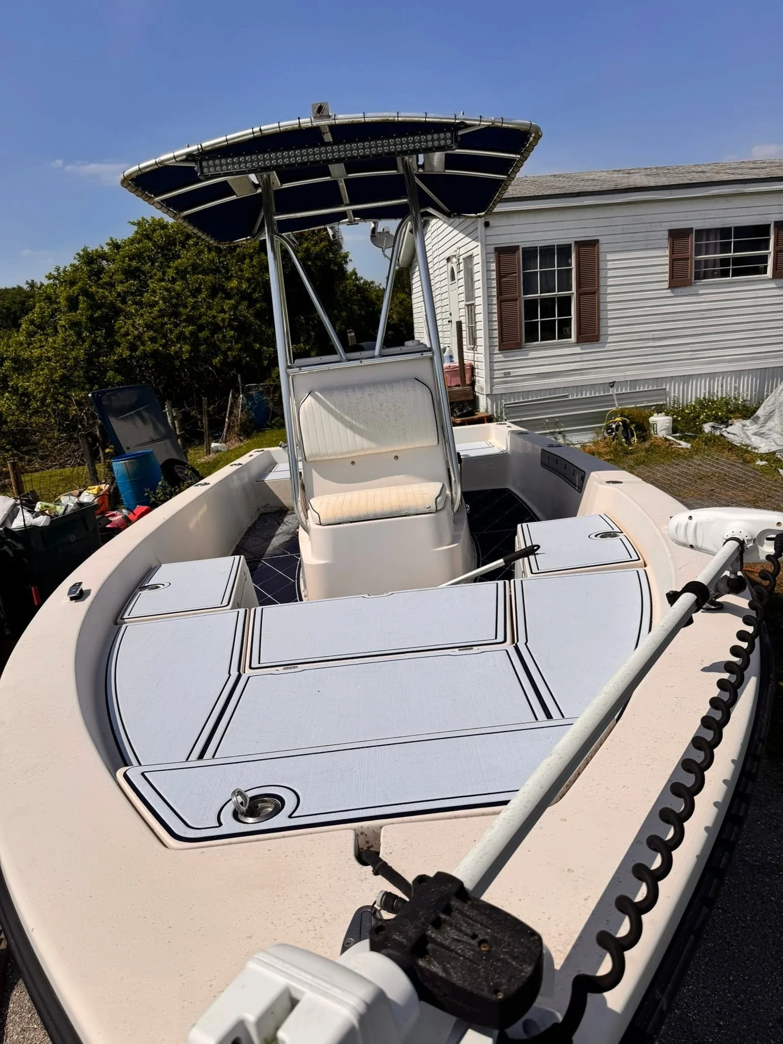 The image shows a white boat with a tower and a shade canopy, parked in a backyard during daytime, with a house and greenery in the background.