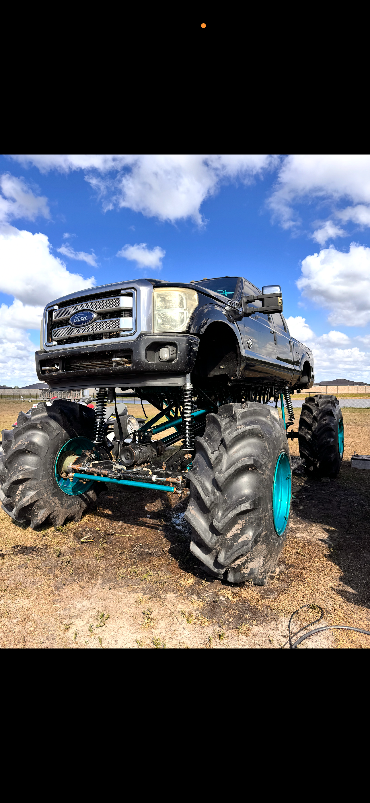 A large monster truck with oversized tires and a black Ford pickup truck body on top, parked outdoors under a blue sky with scattered clouds.