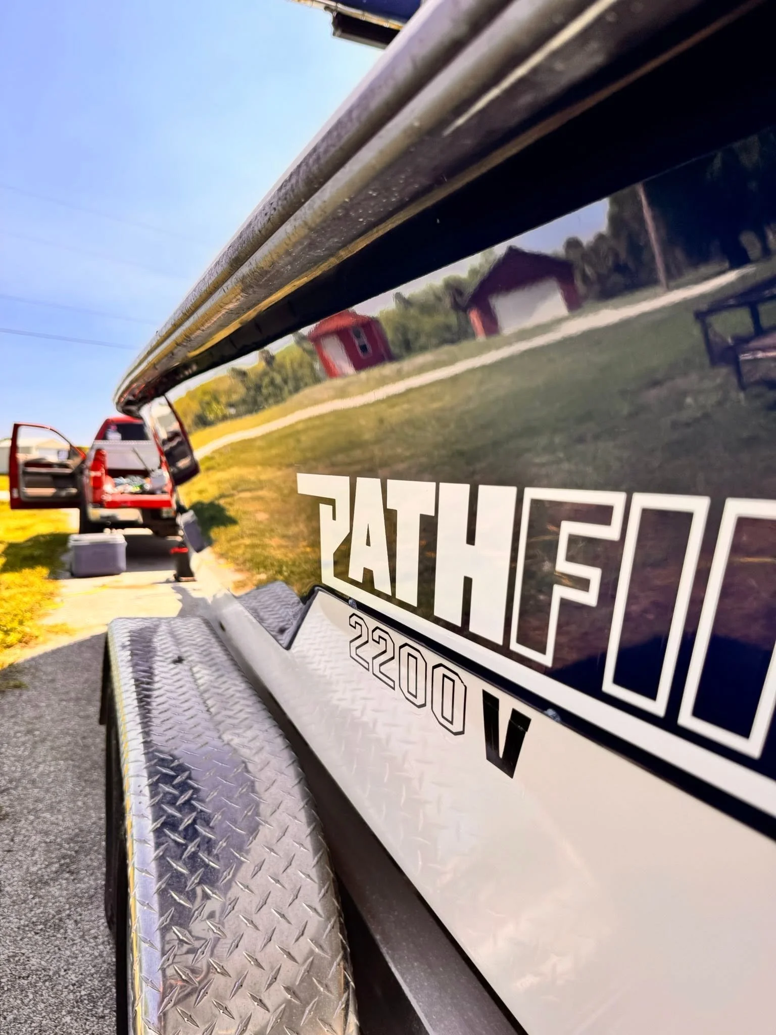 Close-up of a fire truck with a reflective black and yellow surface, showing part of a registration number and a diamond-plate metal step, with a scene of a grassy area, trees, and small red buildings in the background under a clear blue sky.