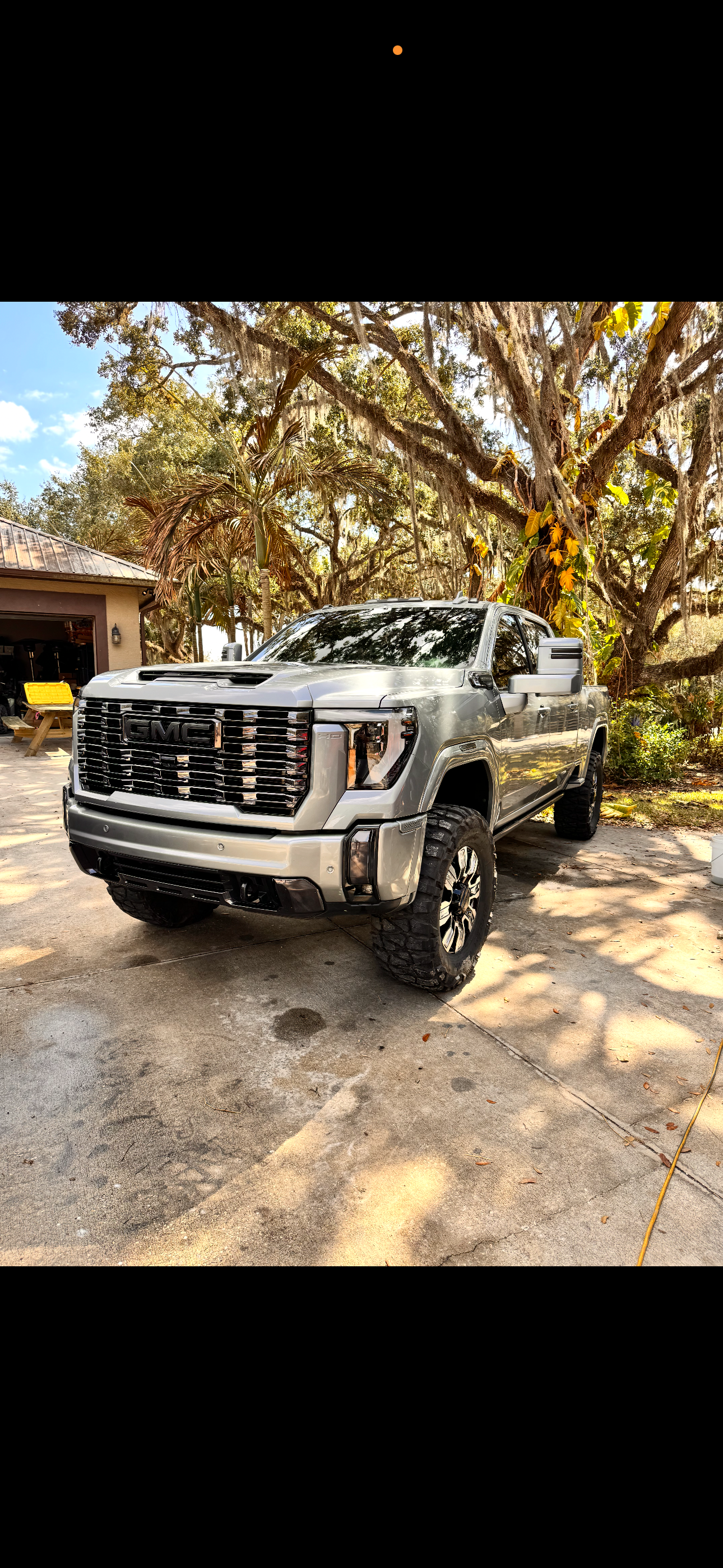 A silver GMC pickup truck parked on a concrete driveway under trees with Spanish moss and leaves, with a clear blue sky in the background.