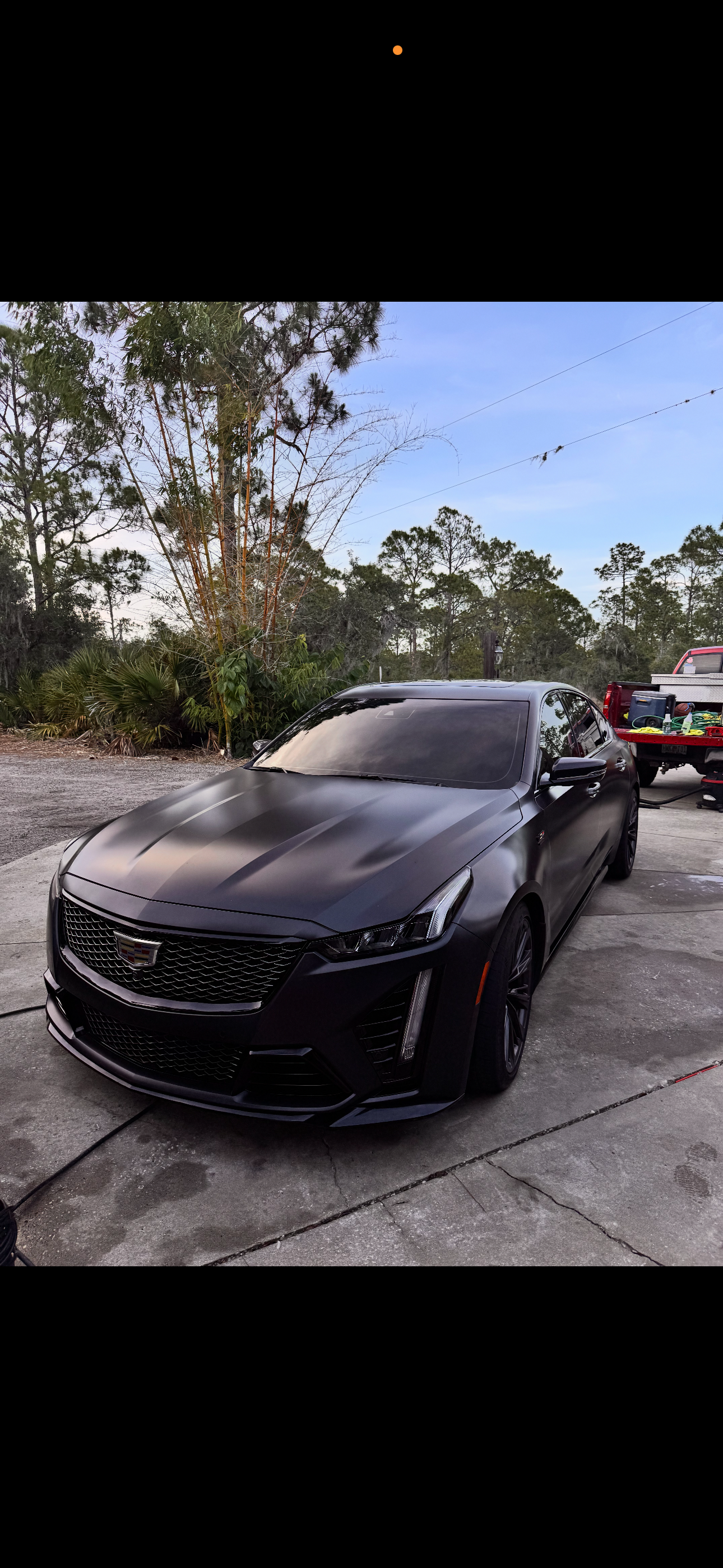 A black Cadillac sports car parked on a concrete driveway, with trees and blue sky in the background, and a red truck with tools in the back nearby.