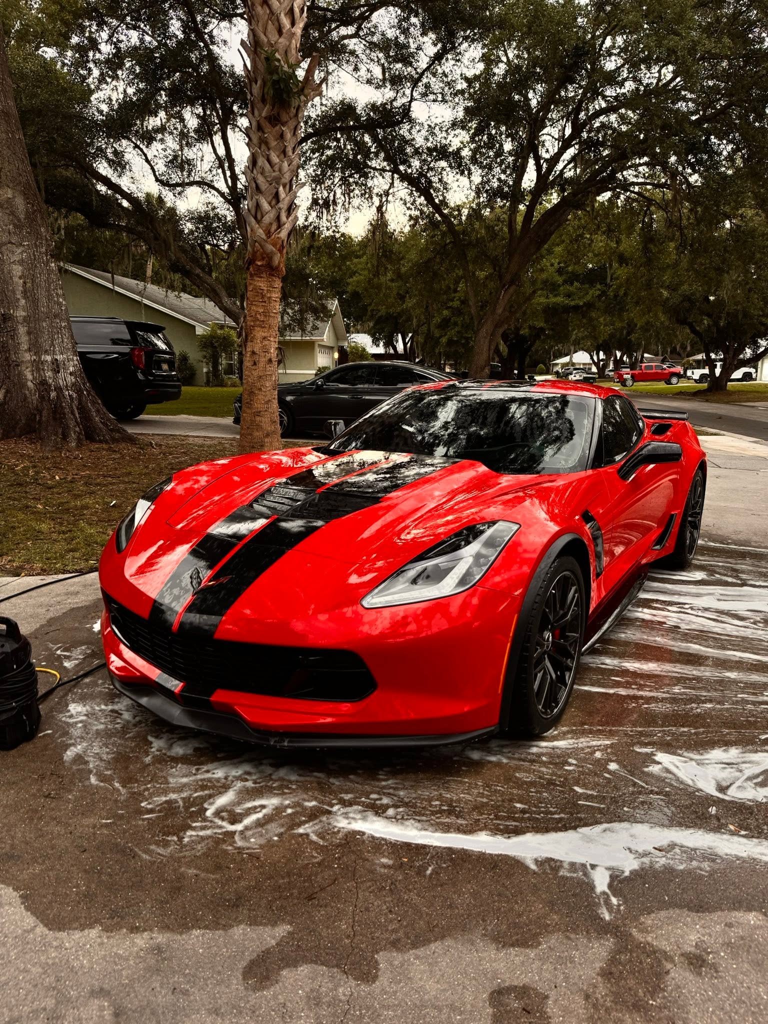 Red sports car with black racing stripe parked next to a house under large trees, with soap suds on the ground from washing the car.
