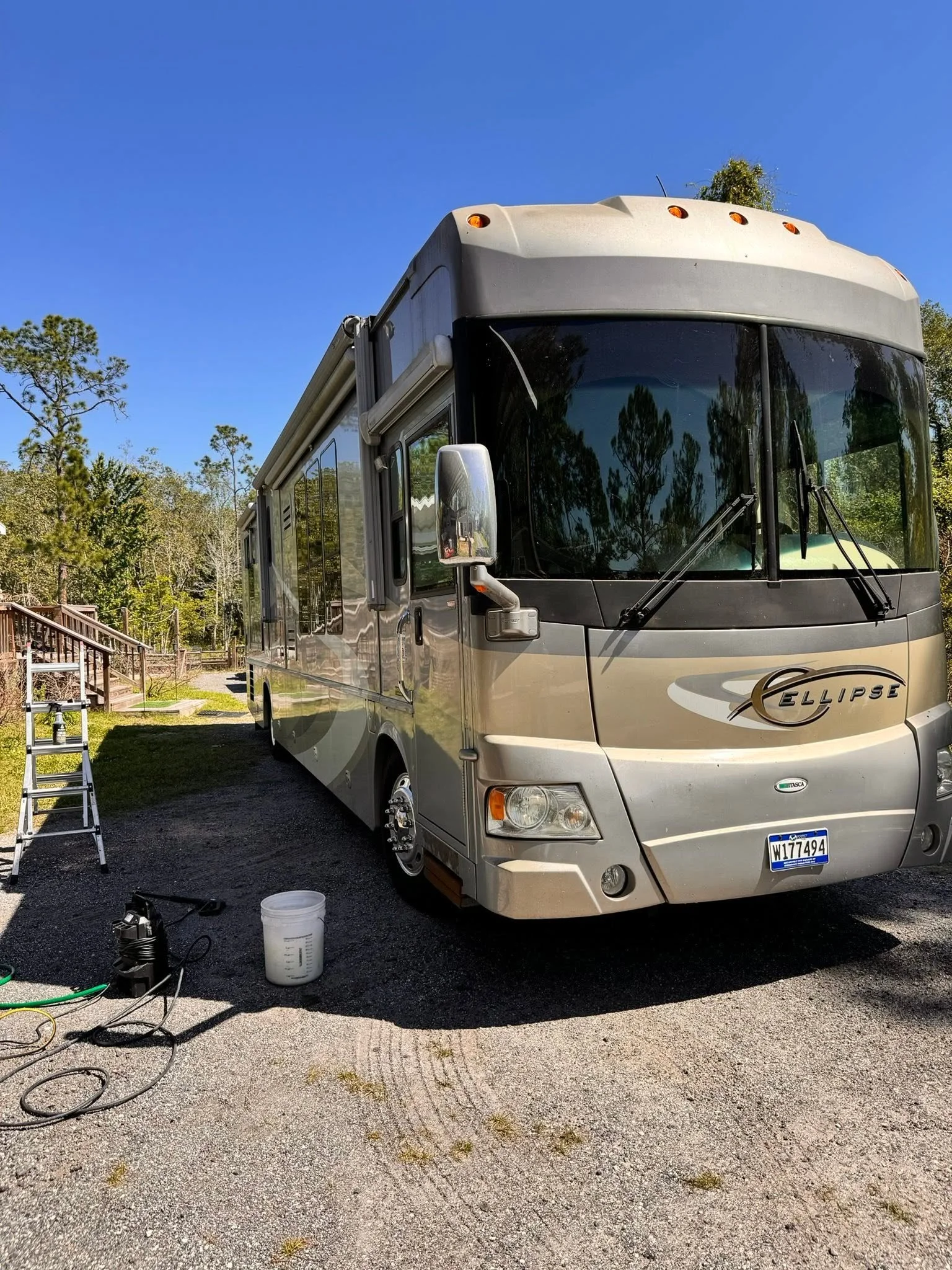 Motorhome parked outdoors with equipment like a ladder, hoses, and buckets surrounding it, and trees in the background under a clear blue sky.