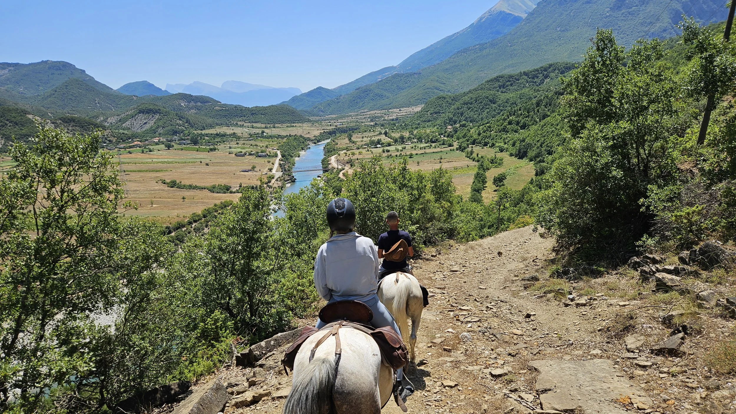 Two people riding horses down a mountain trail overlooking a green valley with a river, fields, and mountains in the background.