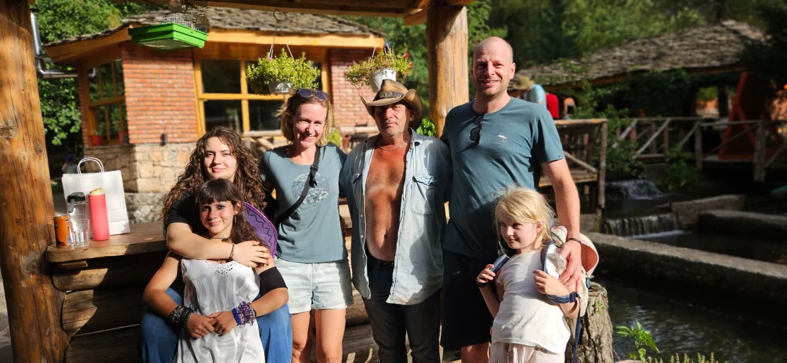 A group of six people, two children and four adults, standing outdoors near a small waterfall and wooden structures, enjoying a sunny day.