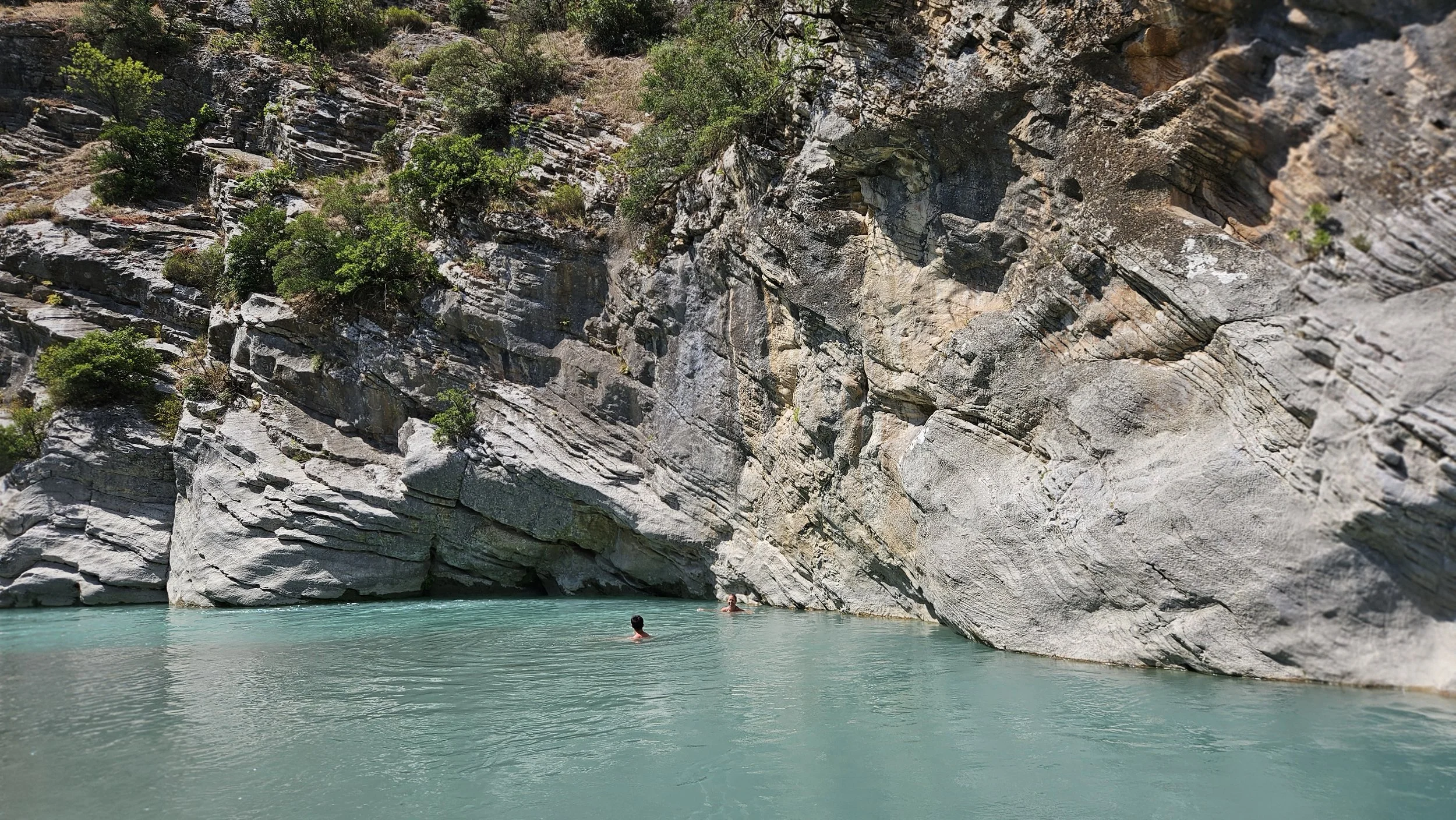 People swimming and relaxing in a turquoise river surrounded by large, rocky cliffs with green vegetation.