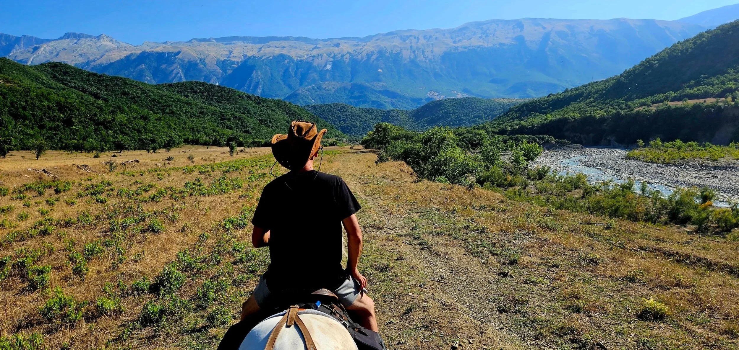 A person riding a horse through a grassy valley with mountains in the background and a river on the right side.