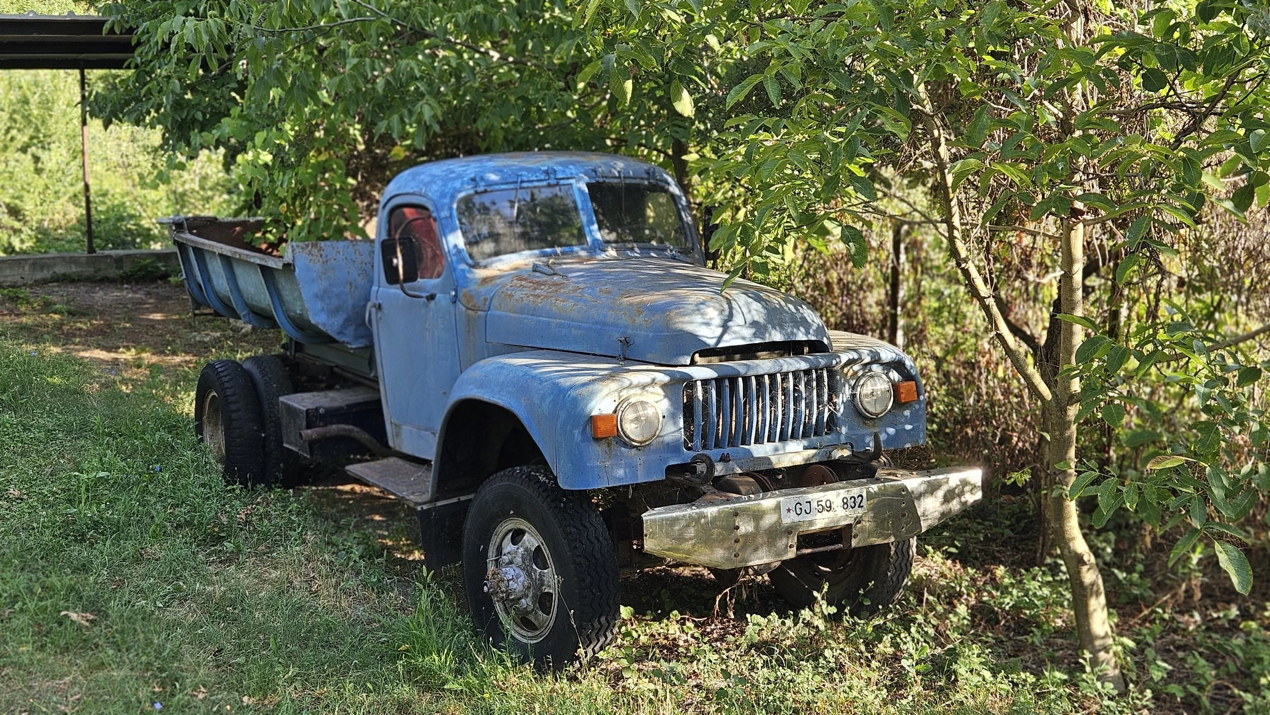 An old, rusted blue truck parked in a garden area surrounded by green trees and grass.