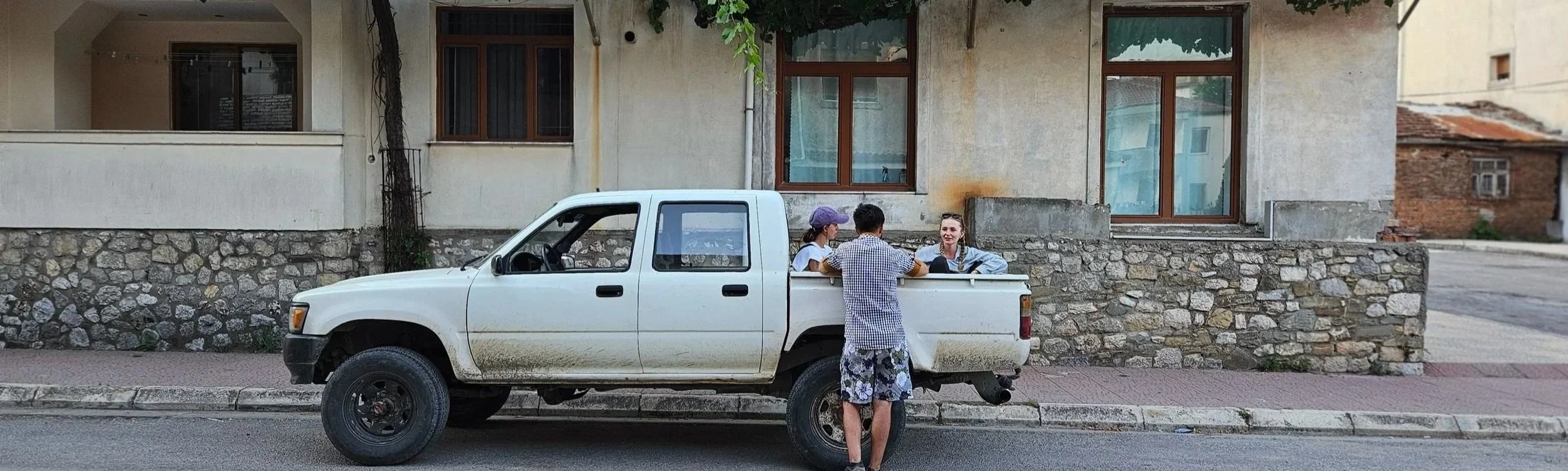 Four people talking next to a white pickup truck parked on the street in front of a building with stone and concrete walls and large windows.