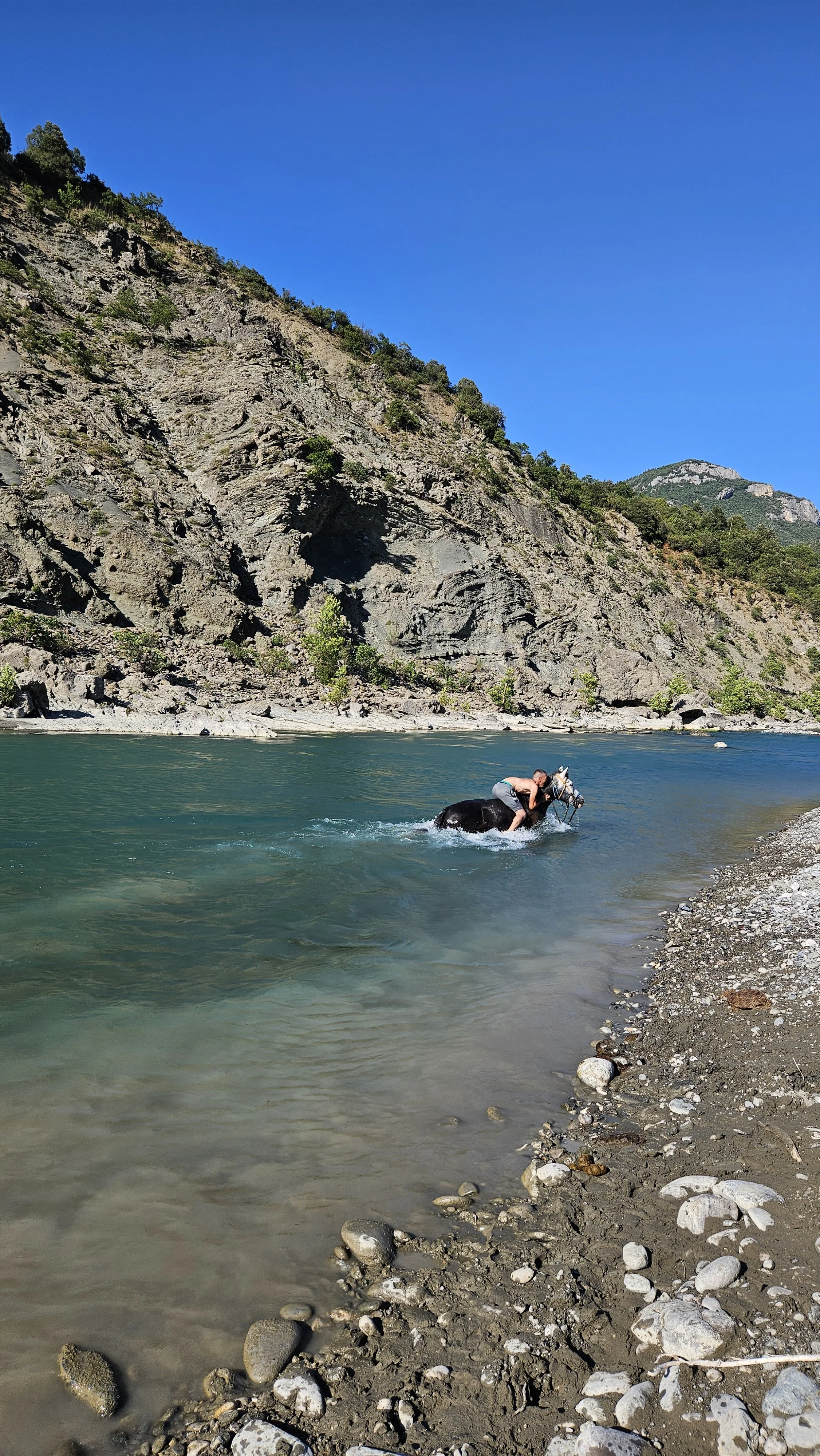 Person riding a black horse in a river surrounded by rocky shoreline and steep mountain cliffs under a clear blue sky.