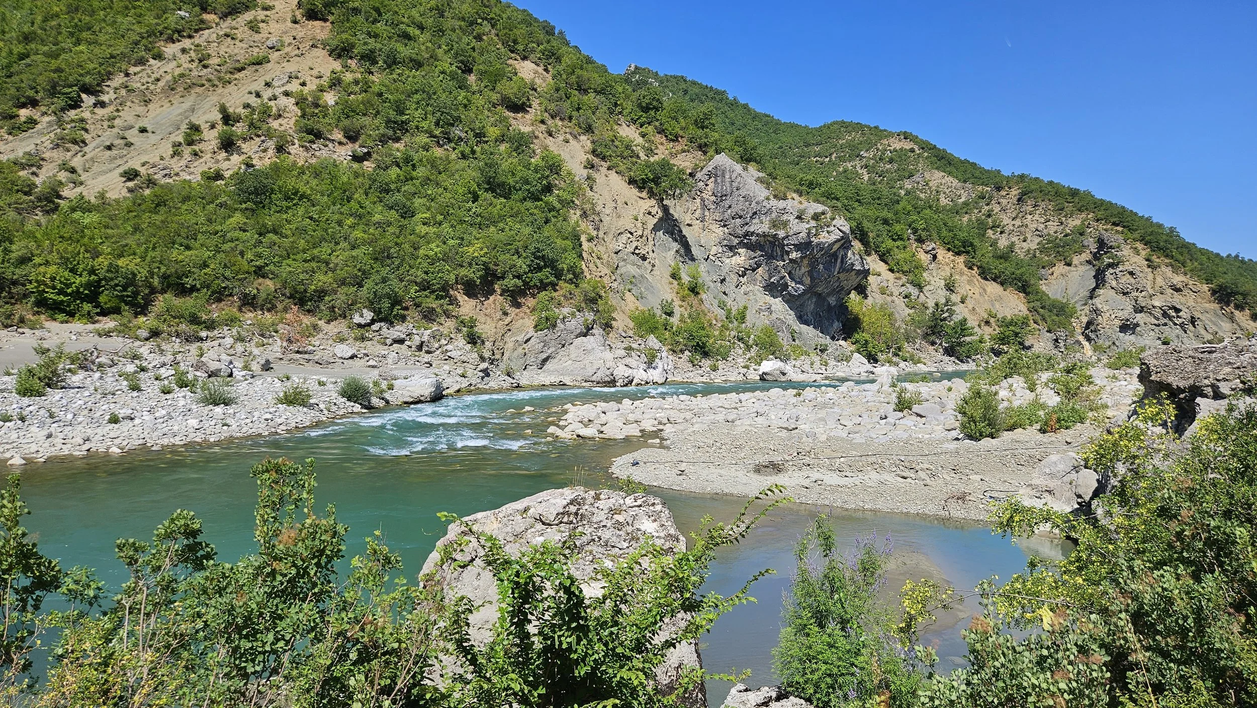 A river flowing through a rocky landscape with green bushes and trees on the banks, and a steep hill with forested areas in the background under a bright blue sky.