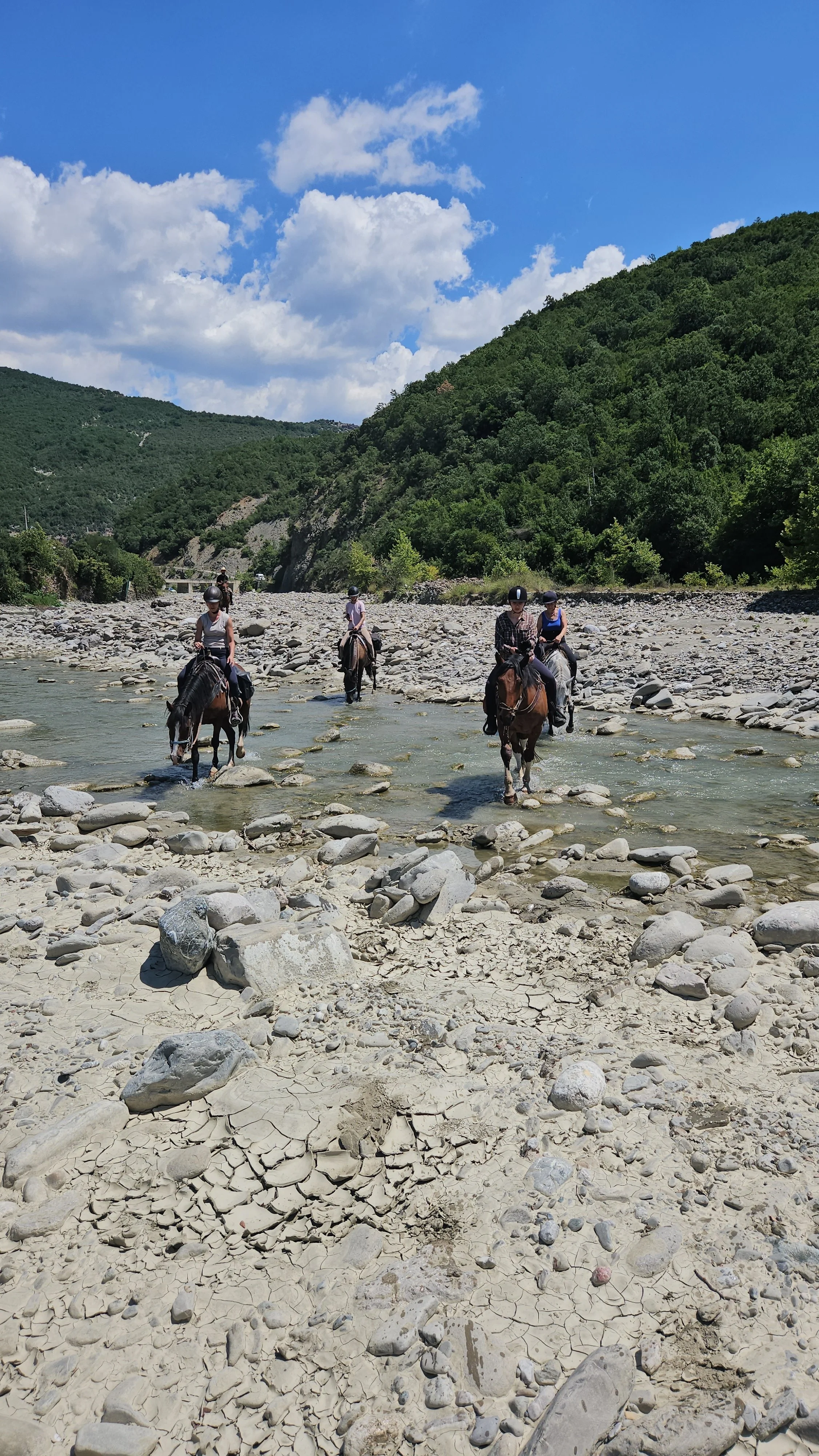 Four people riding horses across a shallow river in a rocky valley surrounded by green hills under a blue sky with clouds.