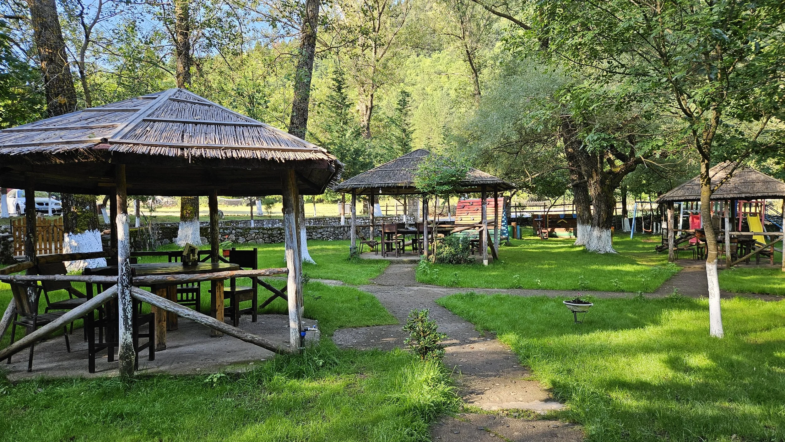 Outdoor park with wooden gazebos, picnic tables, green grass, trees, and a footpath, under a clear blue sky.