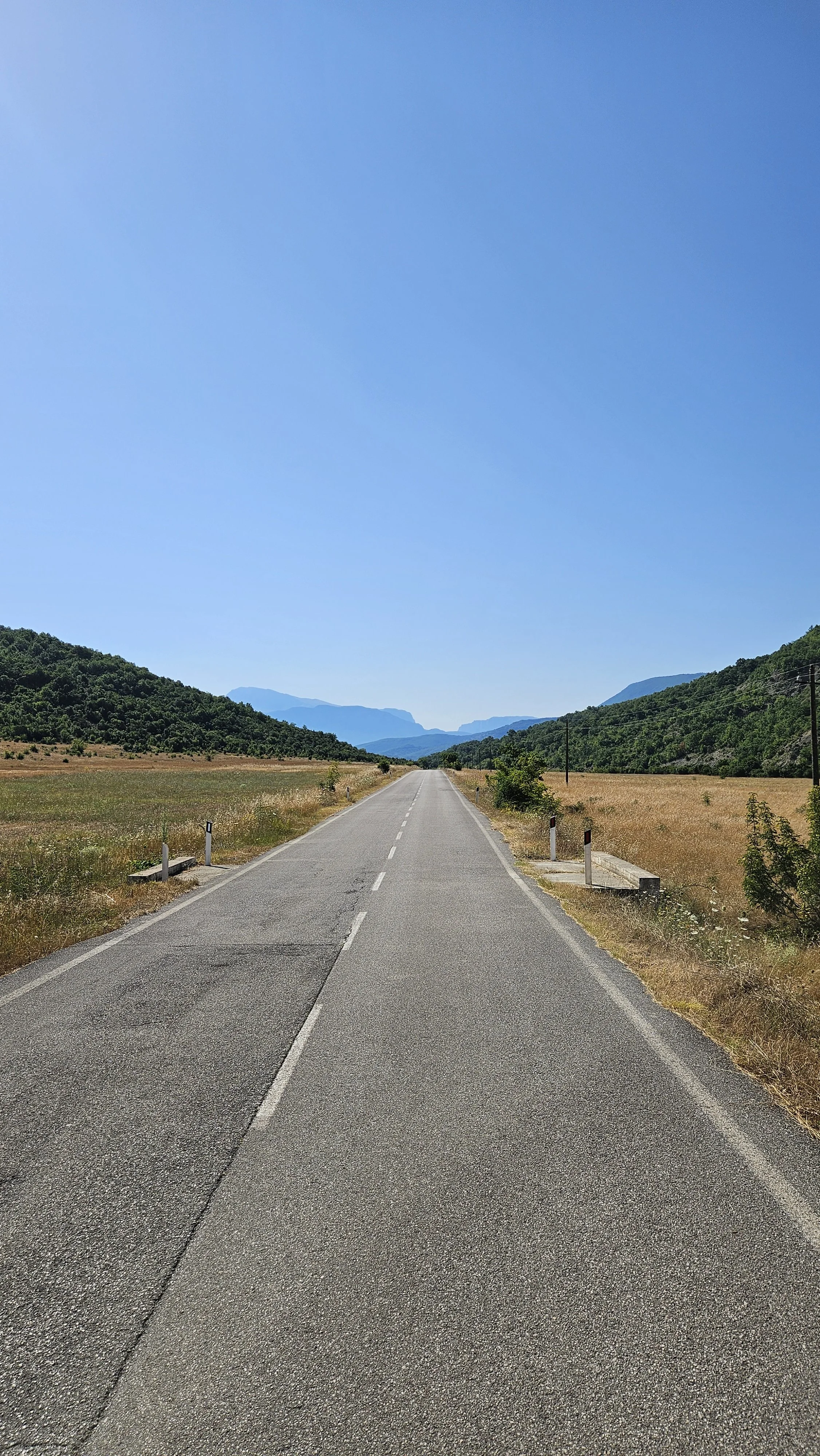 A straight empty road in a rural area with green hills and mountains in the distance, under a clear blue sky.