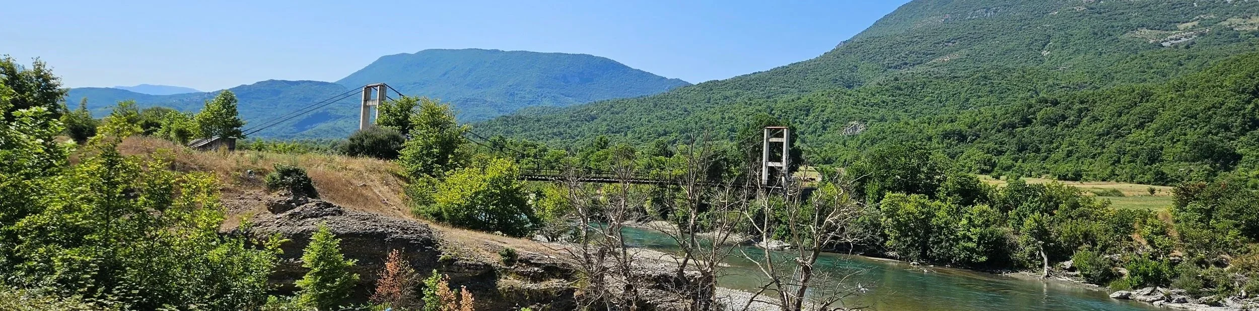 A scenic landscape featuring green mountains, a river, and a small suspension bridge with a single tower, surrounded by dense trees and vegetation on a clear, sunny day.