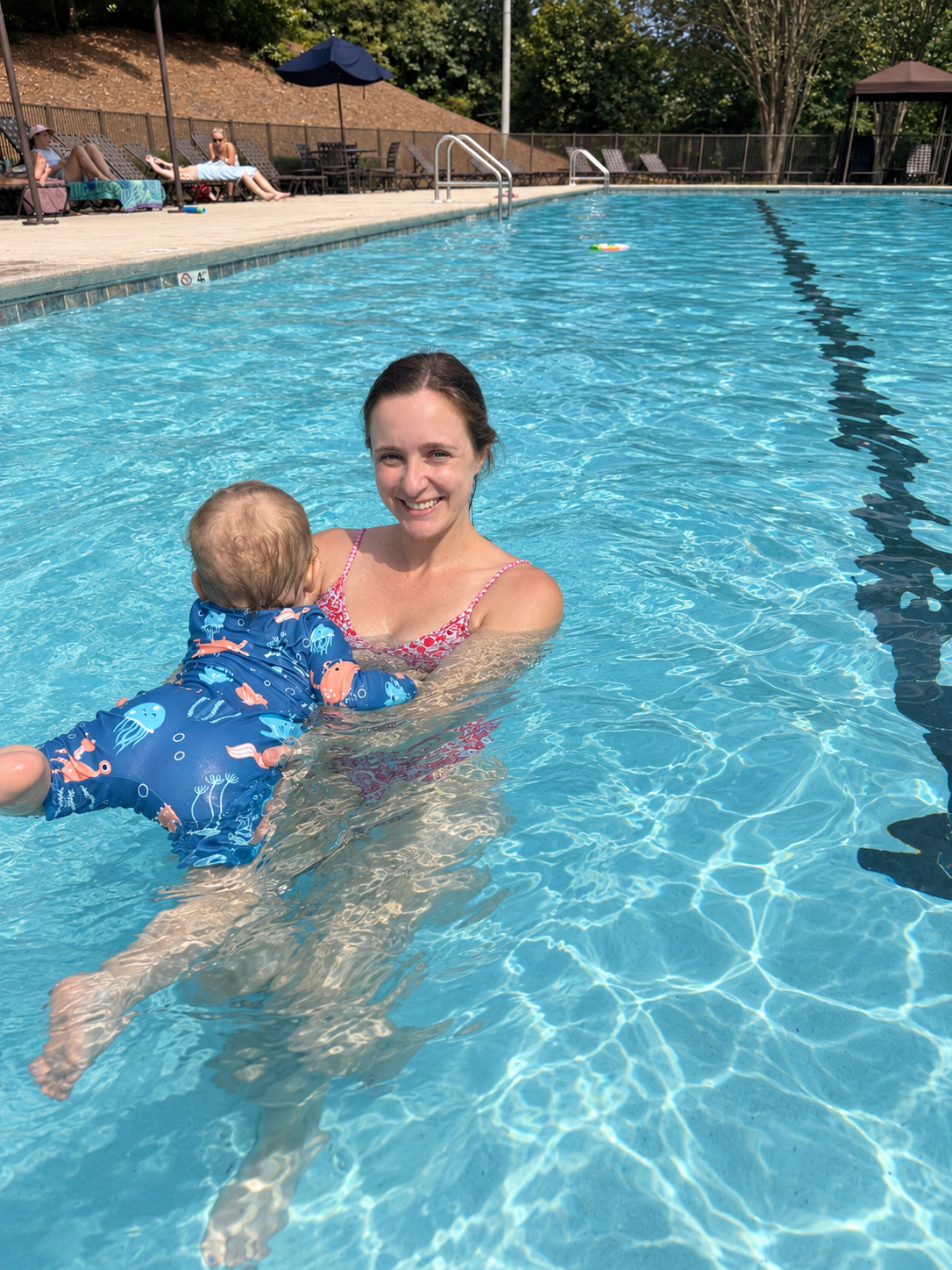 Woman and baby in a swimming pool smiling at the camera, with people relaxing on pool chairs in the background.